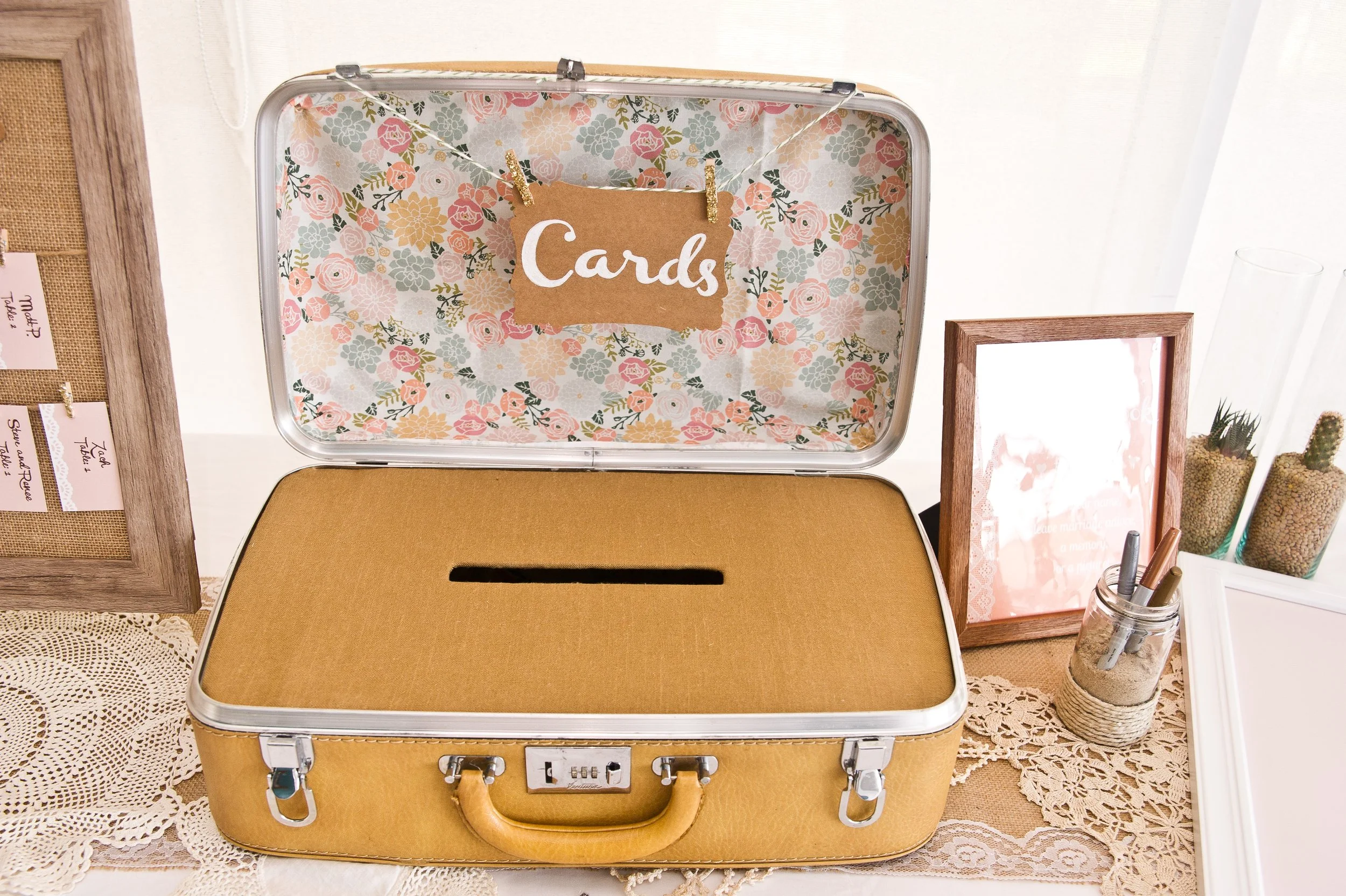 A vintage-style cardboard donation box with a slot on top and a built-in lock is placed on a lace tablecloth, surrounded by framed artwork, decorative plants, and pen holders.