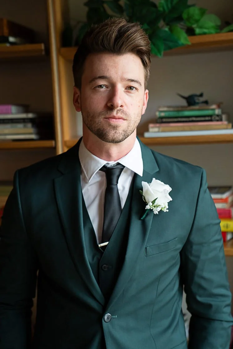 A man in a dark suit with a white shirt, black tie, and boutonnière, standing indoors in front of a wooden bookshelf with books and decorative items.
