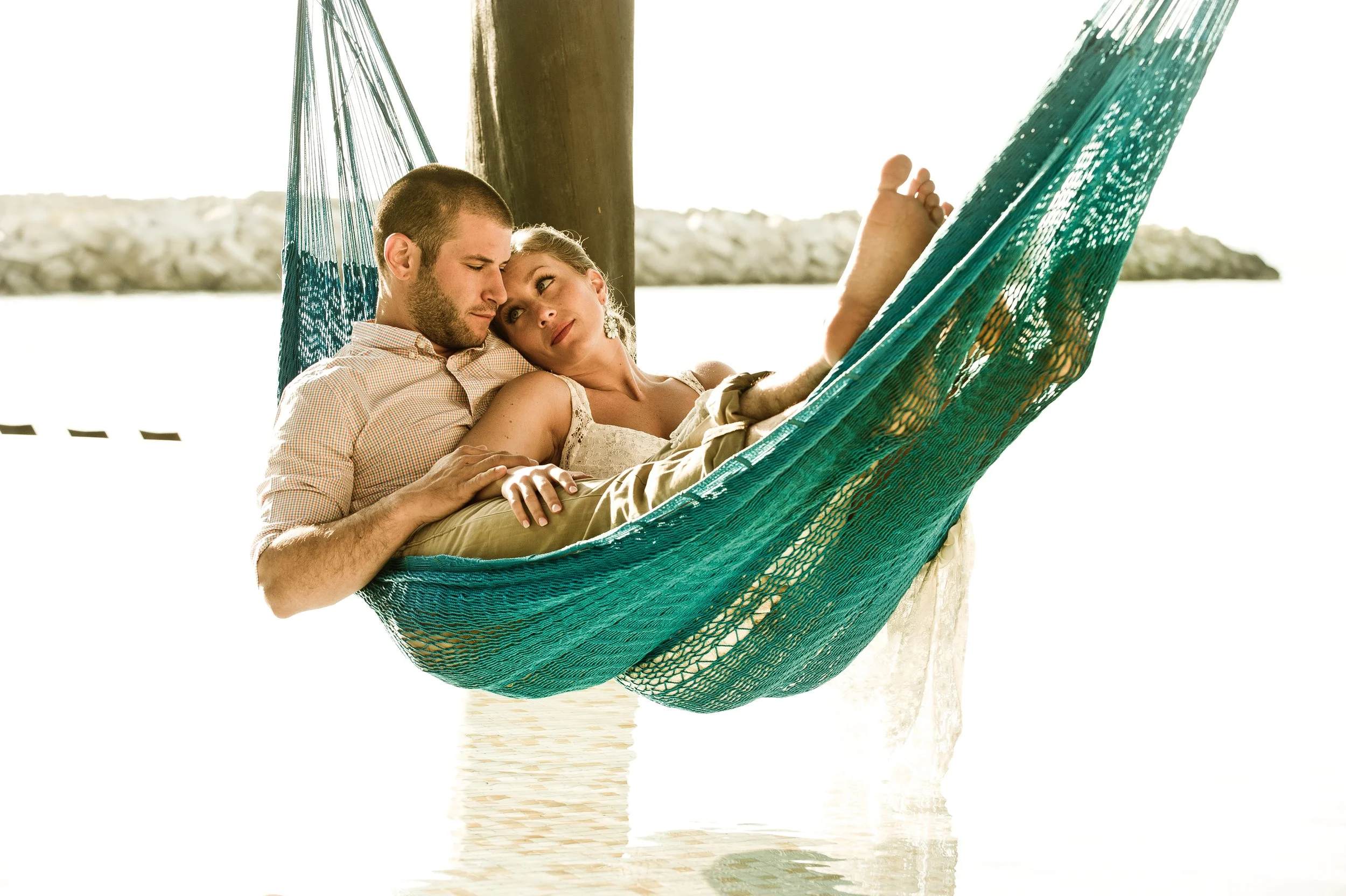 A couple relaxing in a teal hammock by water with rocks in the background.