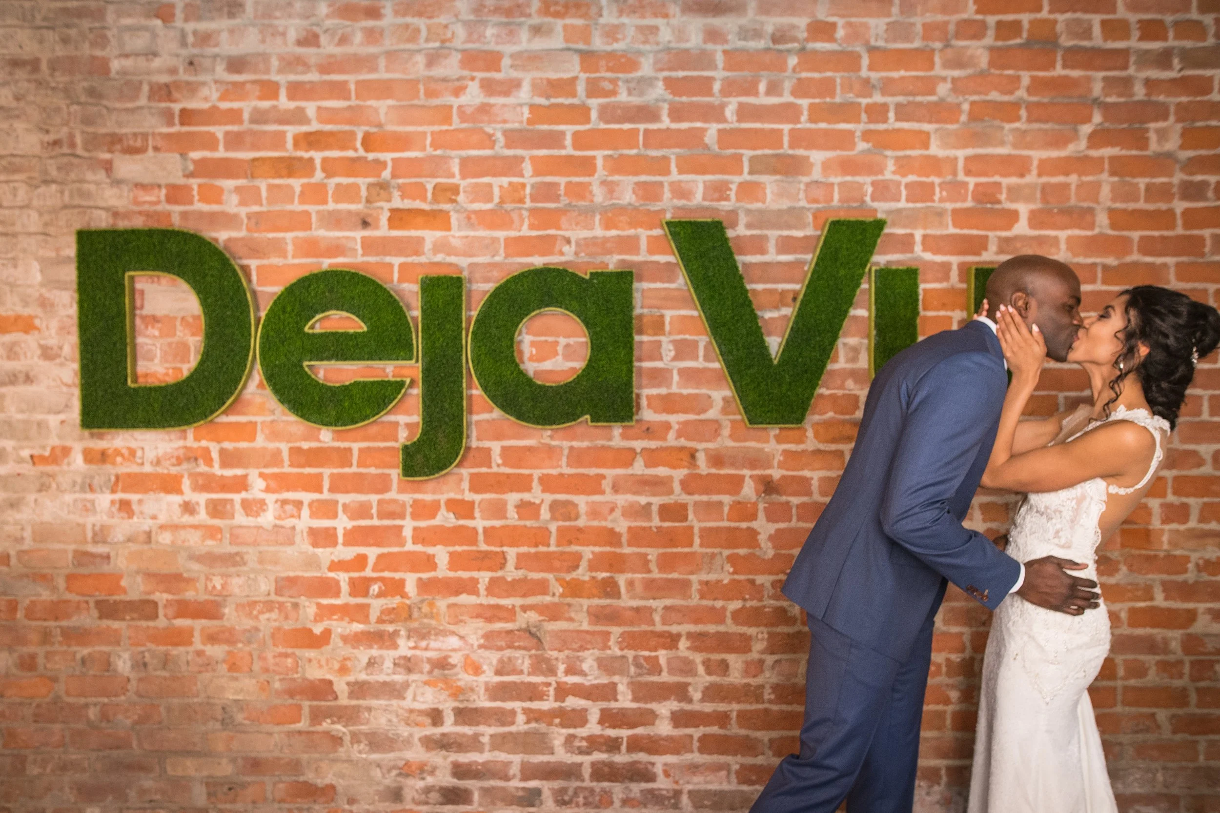 A couple dressed in wedding attire sharing a kiss in front of a brick wall with large green moss letters spelling "DeaV."