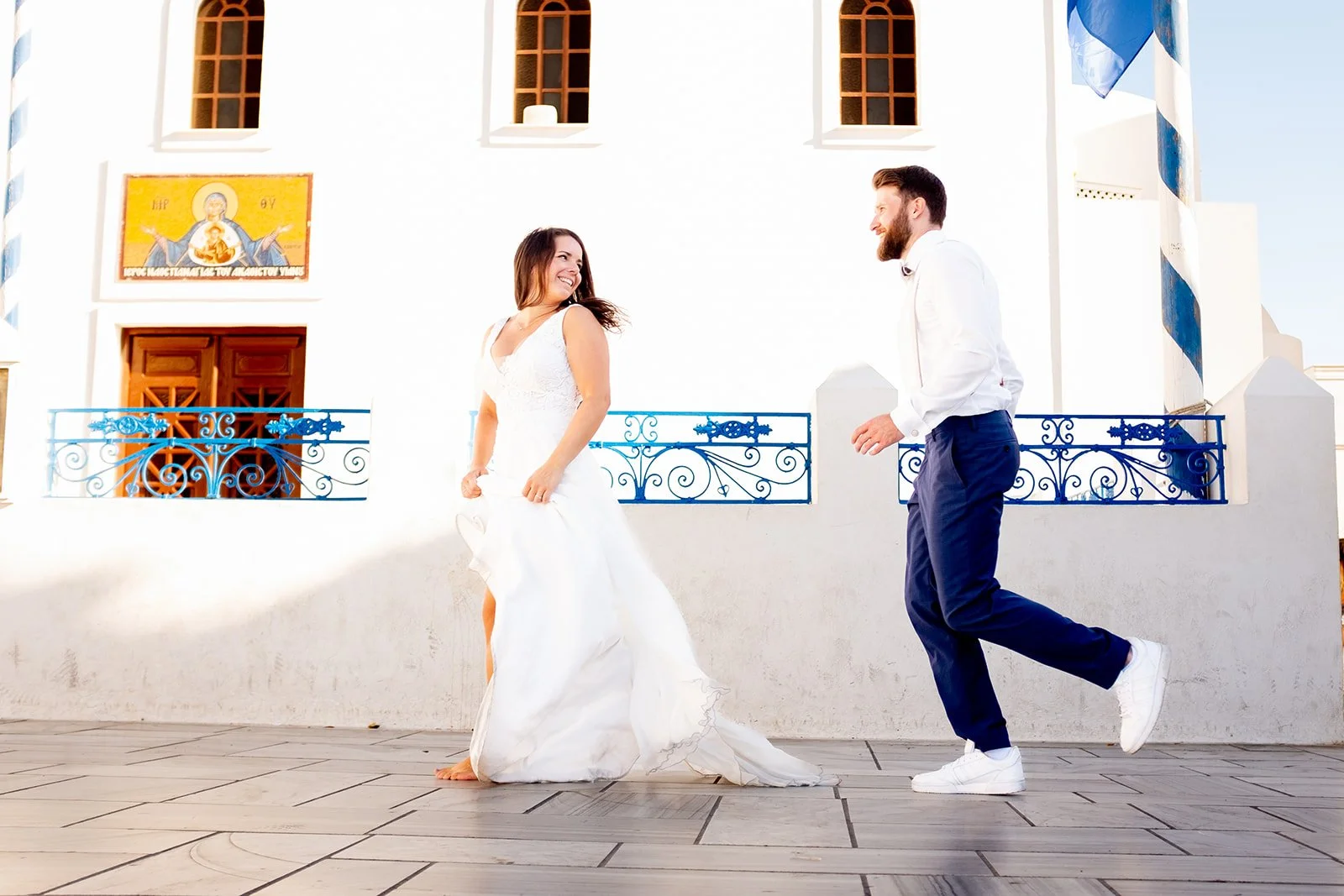 A bride in a white wedding dress and a groom in a white shirt and blue pants dancing outdoors in front of a white building with blue railings and brown windows
