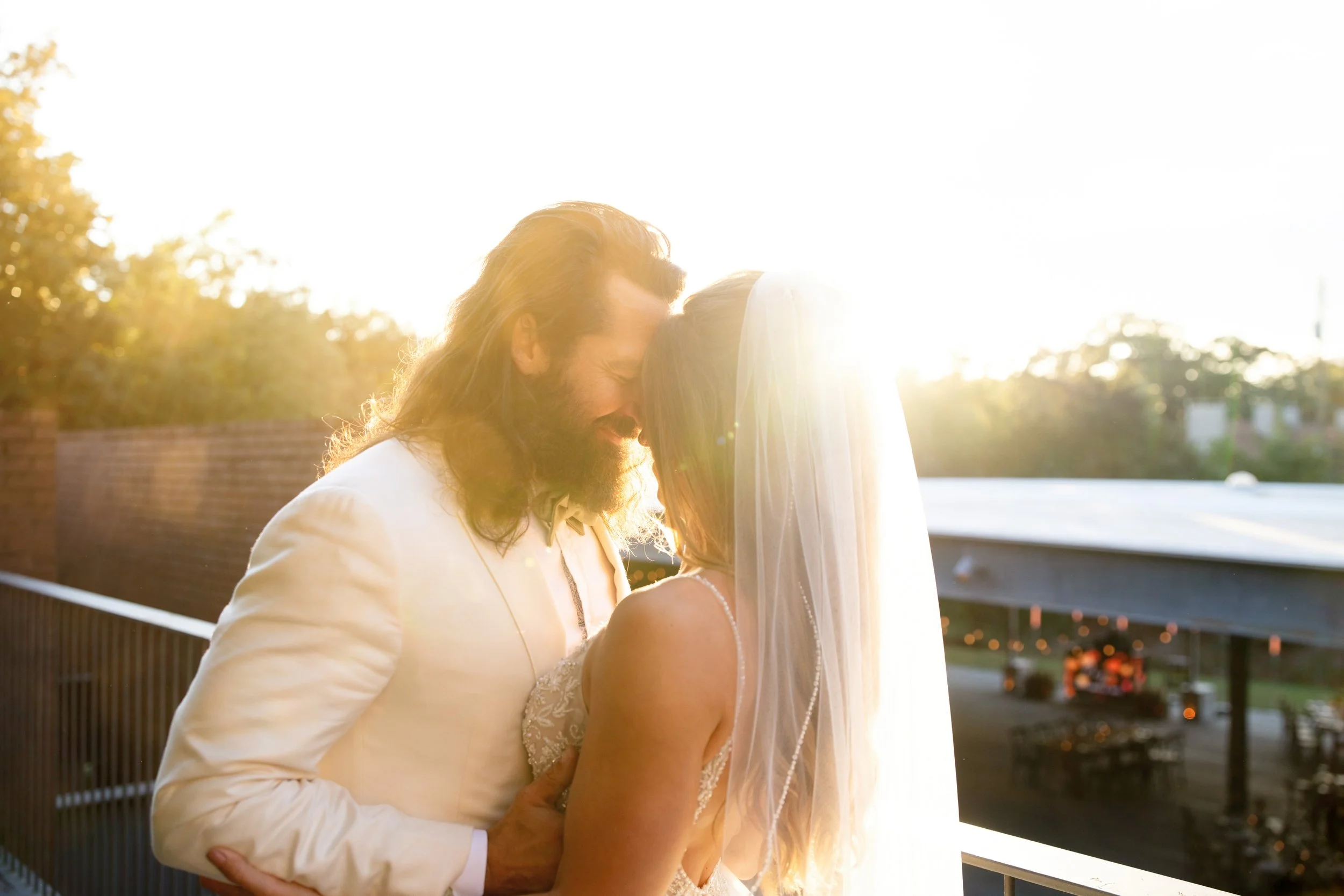 A groom with long hair and a beard, dressed in a white suit, and a bride in a white wedding dress and veil, standing close together on a balcony during sunset, embracing and smiling.