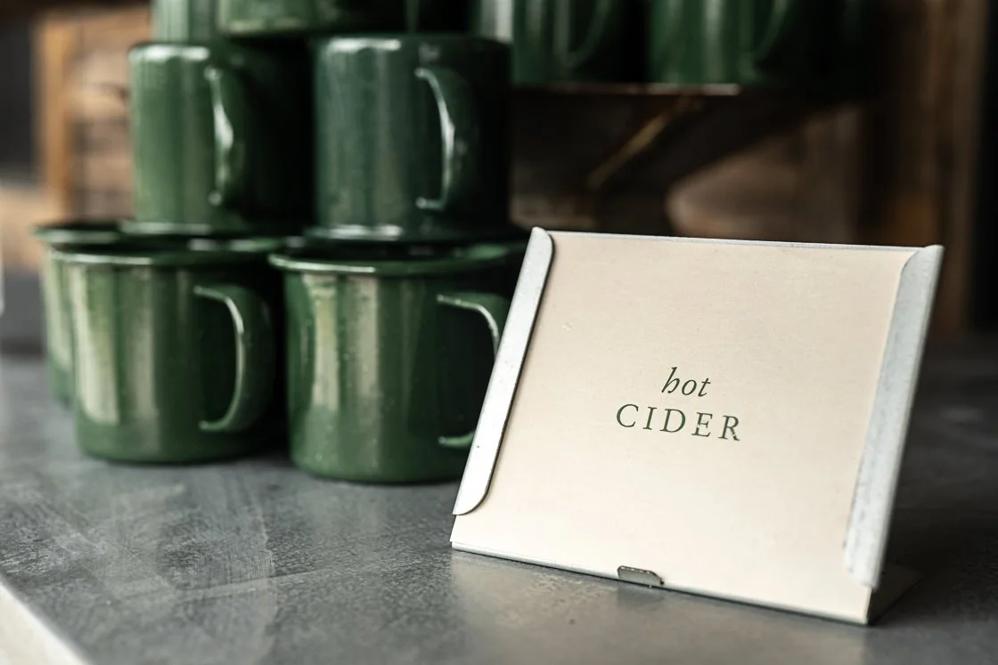 Green ceramic mugs stacked on a dark wooden counter with a small display sign that reads "hot CIDER".