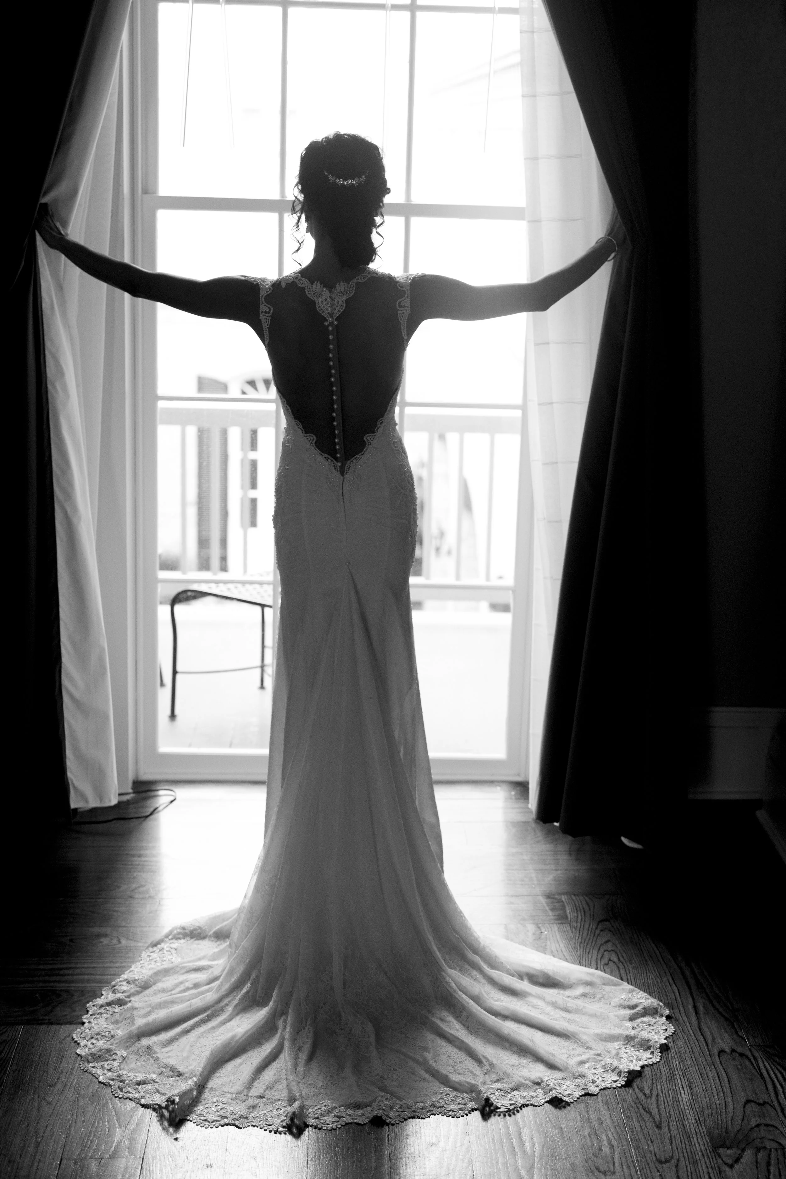A woman wearing a wedding dress stands in front of a large window, opening the curtains to reveal outside light, with her back facing the camera.