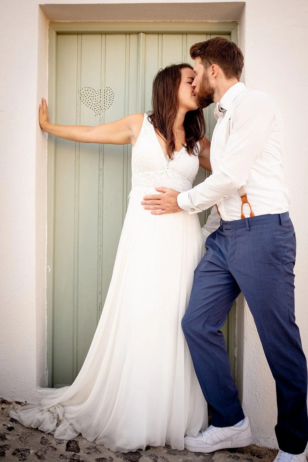 A couple in wedding attire sharing a kiss in front of a green door with a heart cutout. The bride is wearing a white lace wedding dress, and the groom is dressed in a white shirt, navy pants, and suspenders.