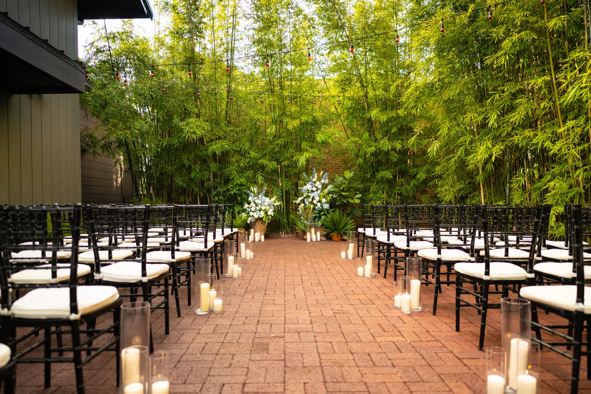 Outdoor wedding ceremony setup with black chairs, white cushions, candles, and floral arrangements under string lights, surrounded by tall green bamboo plants.