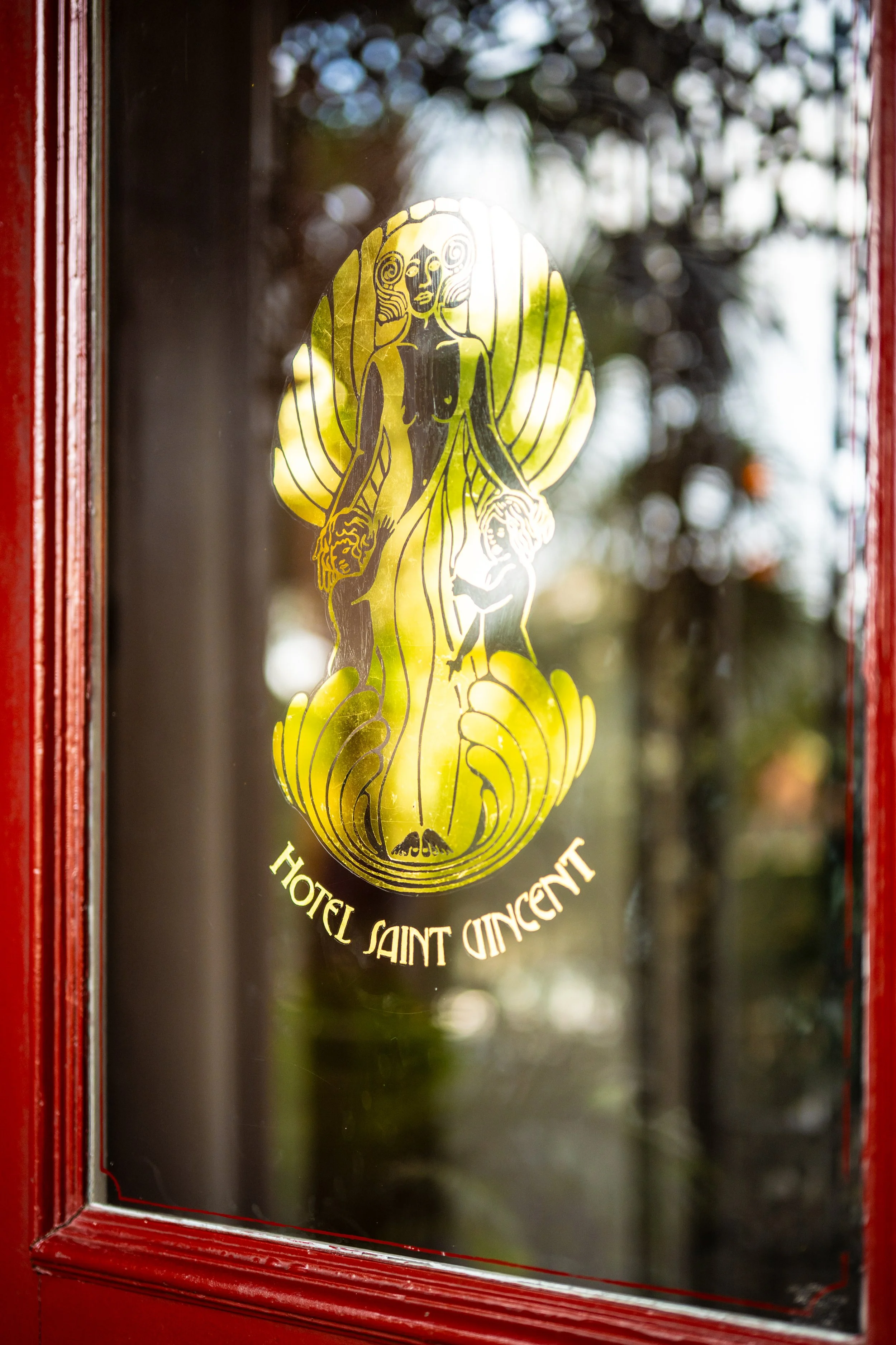 A glass window with a gold-colored logo of Saint Vincent Hotel, depicting a stylized woman with flowing hair, holding two lion heads in her hands. The logo includes the text "Hotel Saint Vincent" at the bottom.