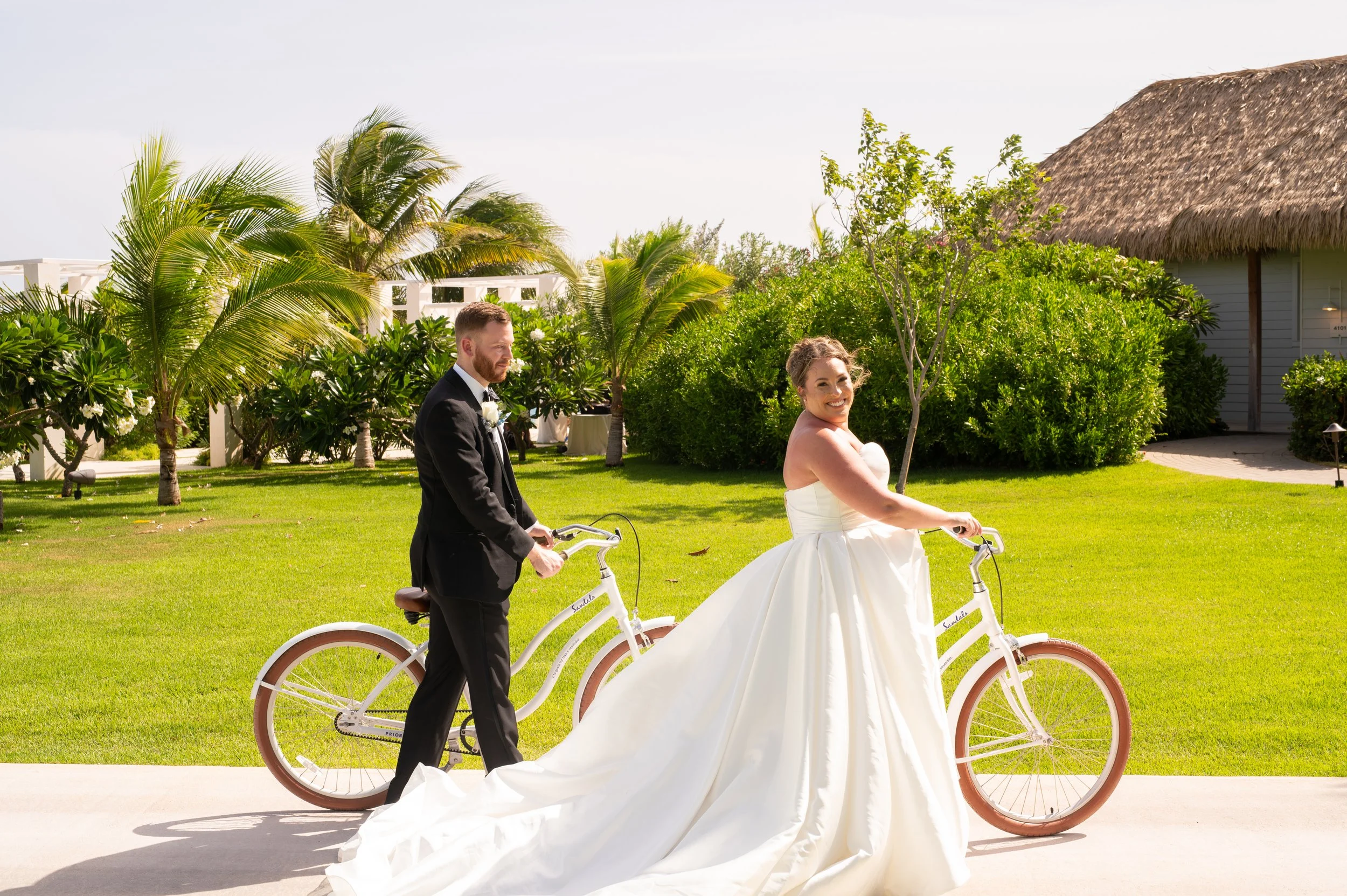 A couple in wedding attire riding white tandem bicycles in a lush green outdoor setting, with tropical trees and a thatched-roof structure in the background.