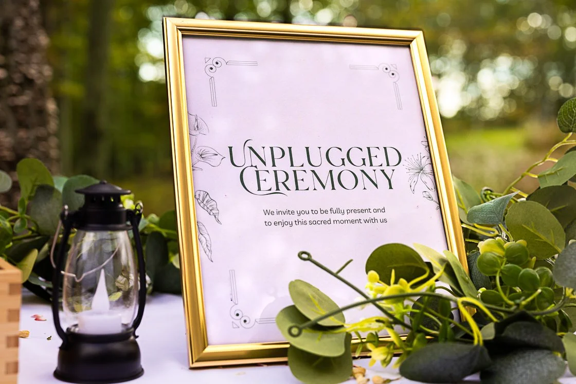 A framed sign titled 'Unplugged Ceremony' on a table decorated with green foliage and a small lantern. The sign invites guests to be present and enjoy a sacred moment.