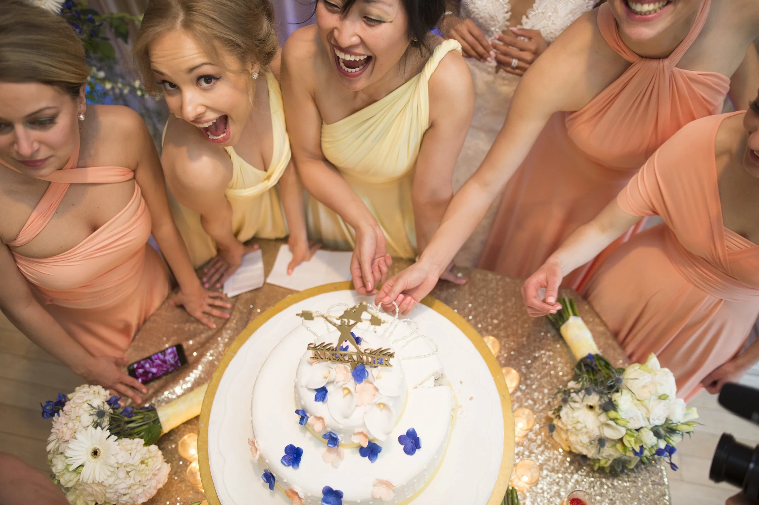 A group of women in pastel dresses gathered around a white cake with flowers and a 'Happy Wedding' topper, celebrating at a wedding reception.