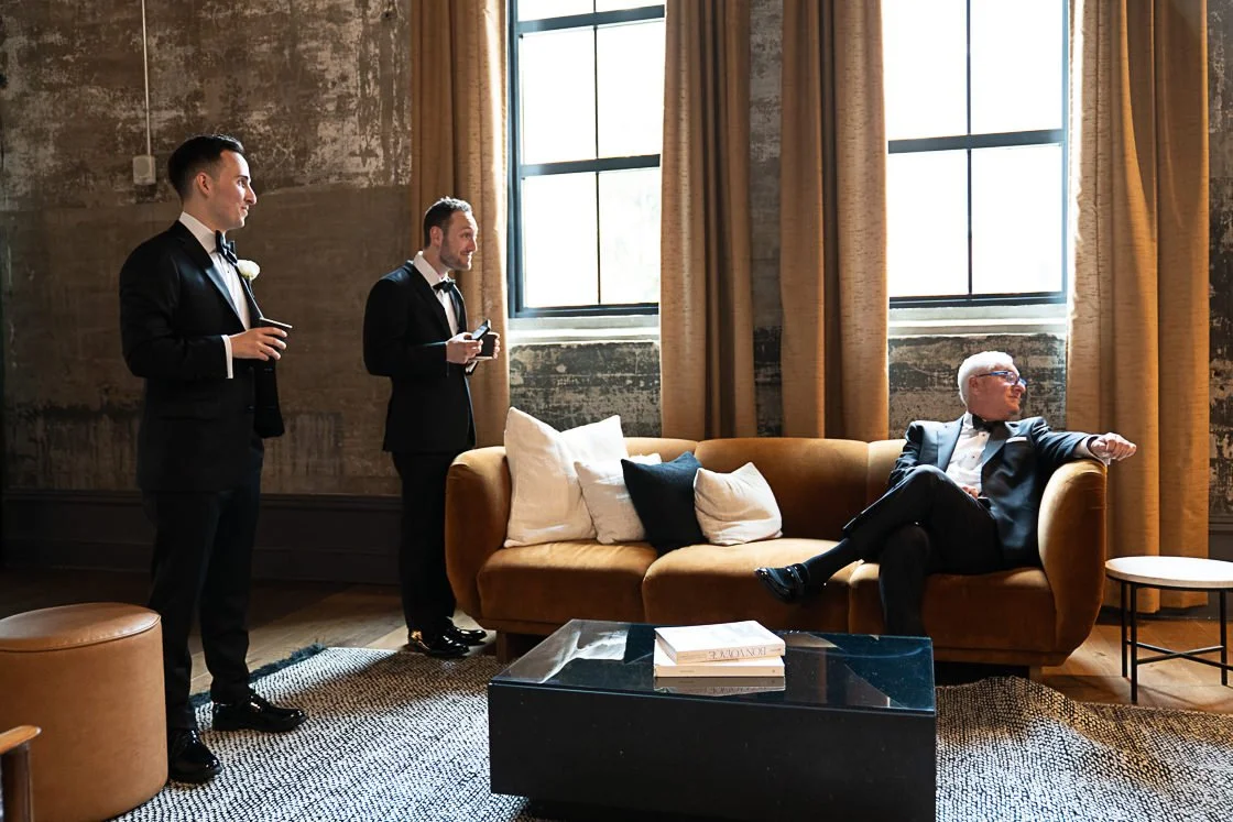 Three men in suits at a social gathering, two standing and one seated on a beige couch, in a room with large windows and curtain, with a black coffee table and decorative pillows.