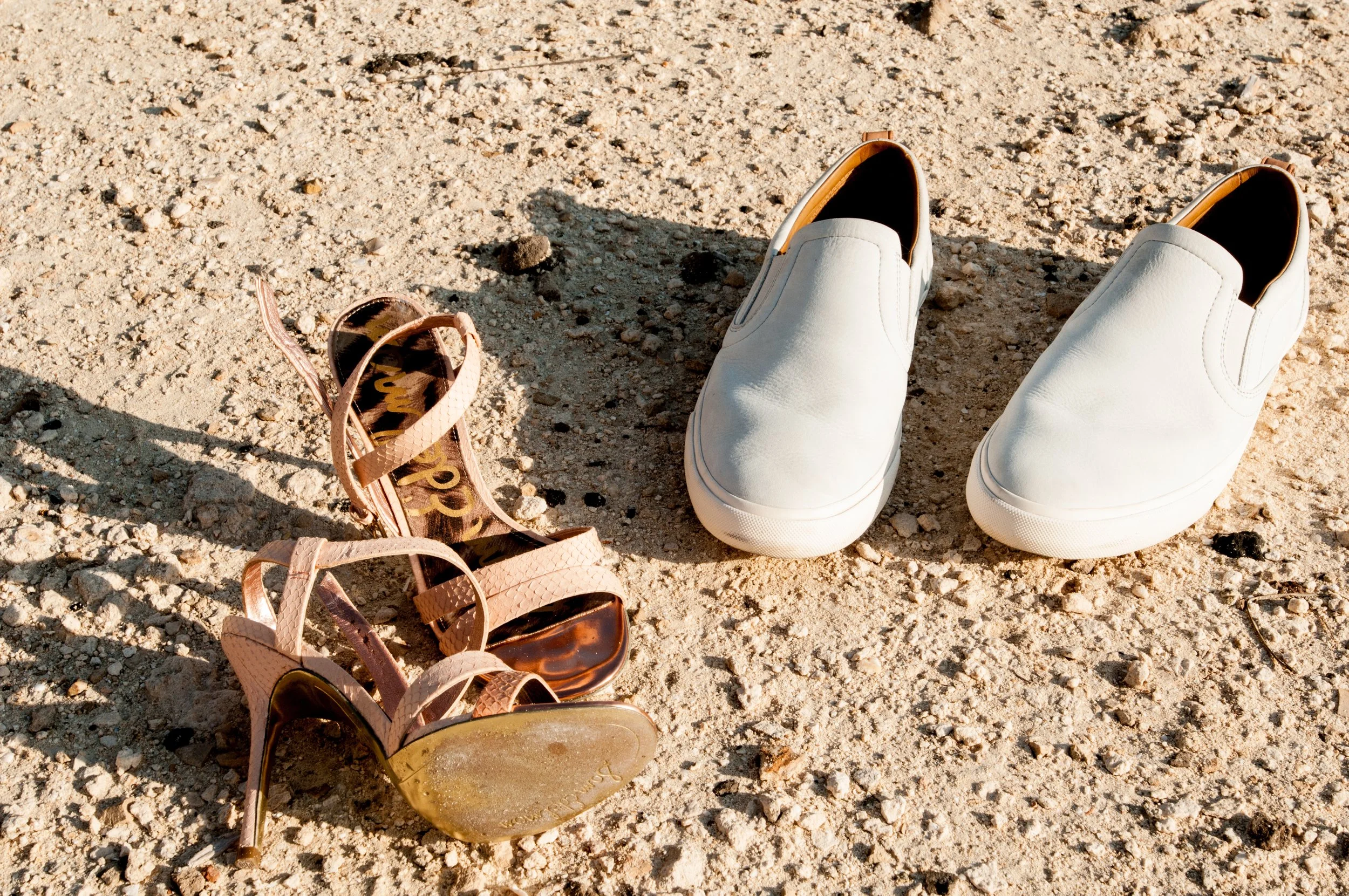 White slip-on shoes and pink sandals with sunglasses on a sandy surface.