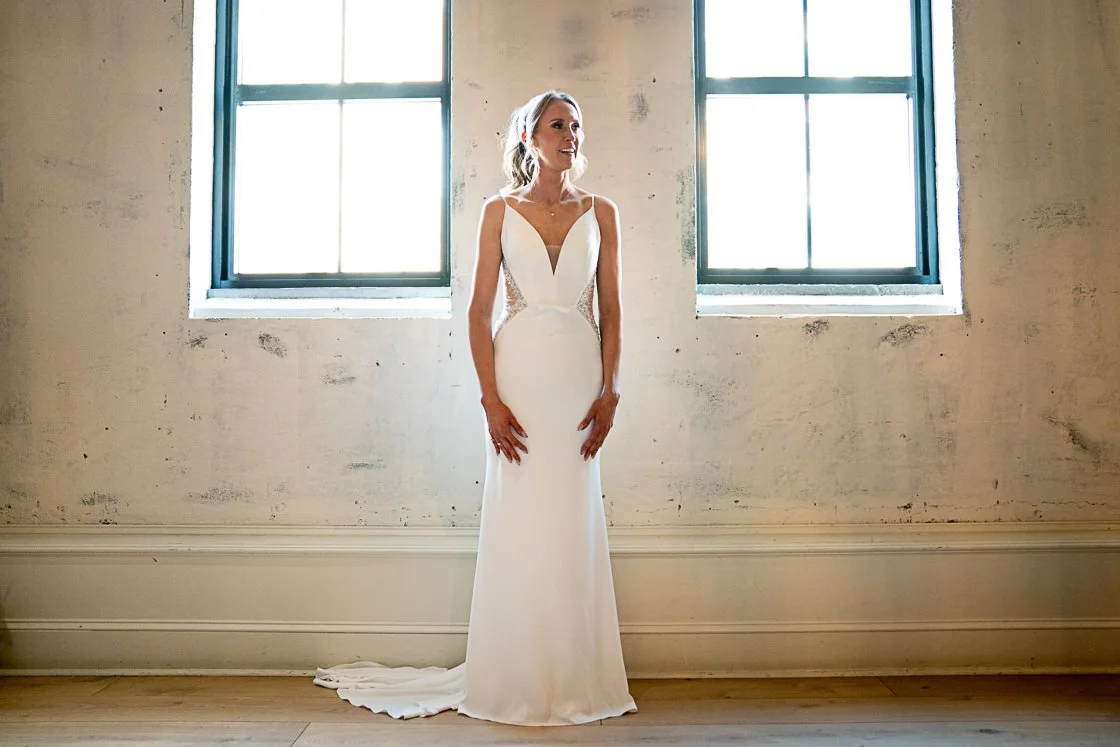 A woman in a white wedding dress standing inside near two large windows with sunlight shining through.