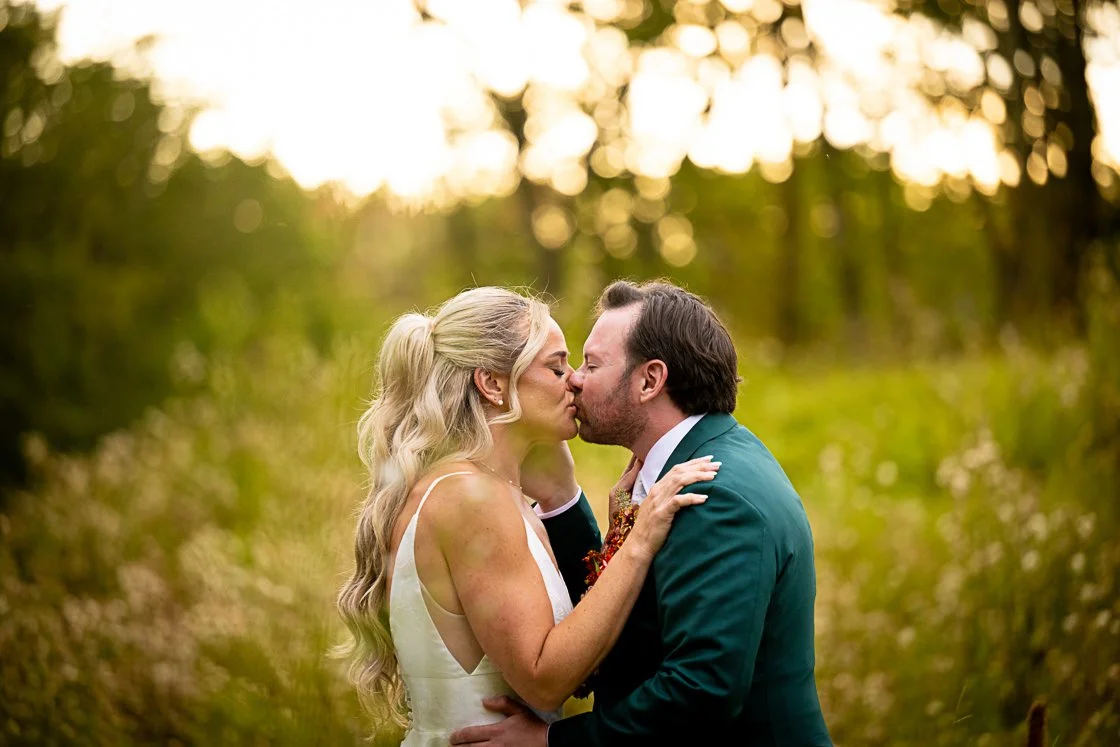 A couple shares a romantic kiss outdoors during sunset, surrounded by trees and greenery.