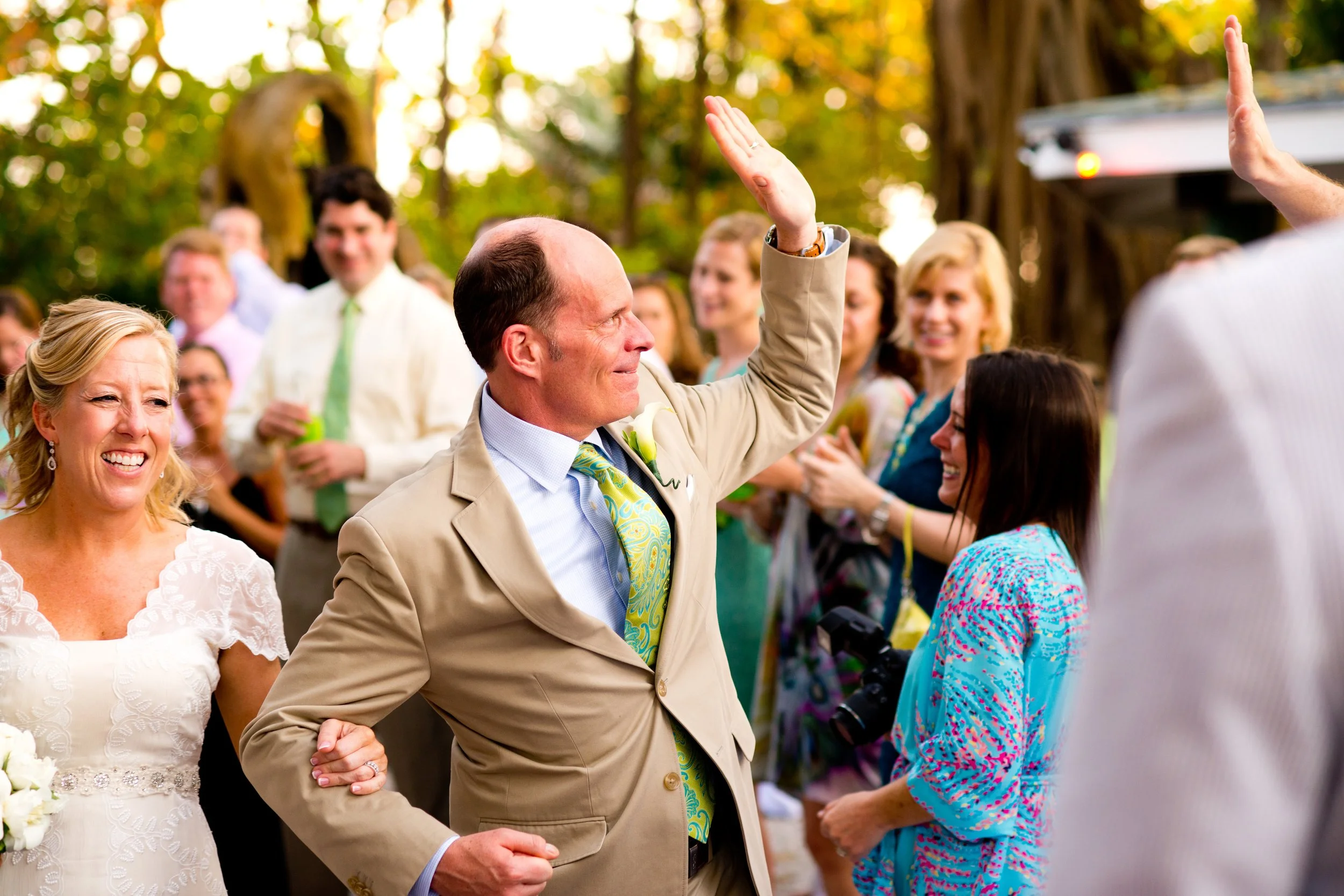 A wedding reception outdoors with multiple guests, a man in a beige suit raising his hand, and women smiling and engaging in conversation, surrounded by trees with golden foliage.