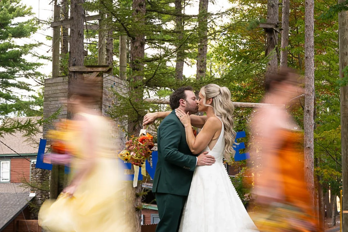 A bride and groom kiss during their outdoor wedding ceremony, surrounded by trees. Two blurred people in colorful dresses are dancing in the foreground, and a wooden sign with blue letters is partially visible behind them.