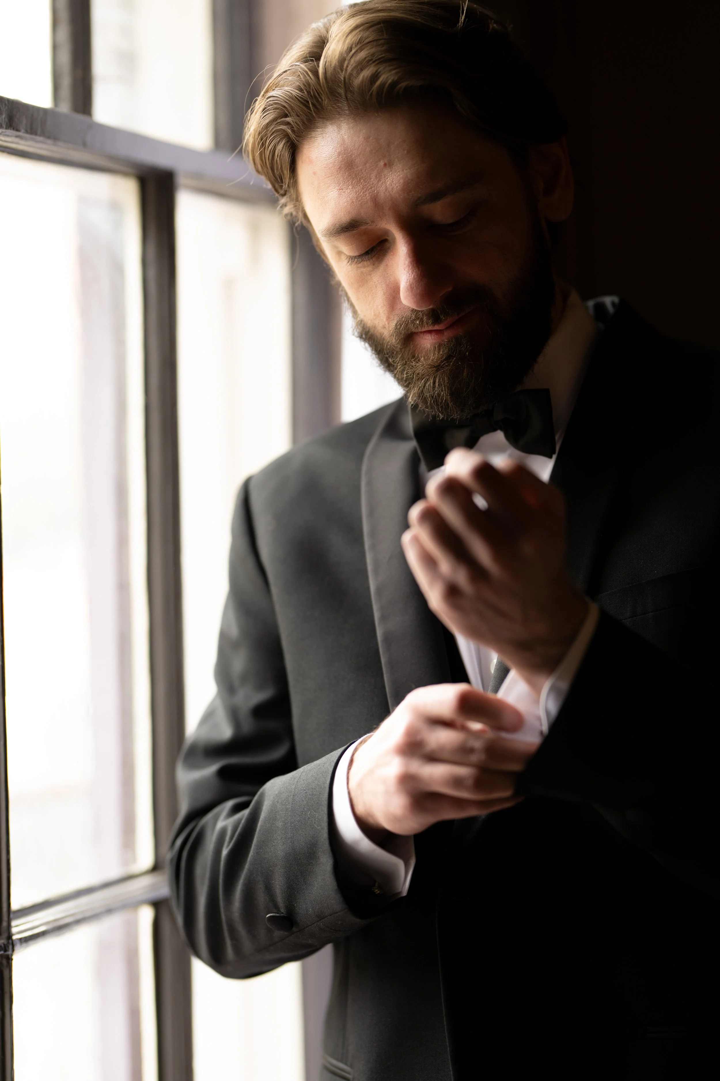 A man in a black tuxedo and bow tie adjusting his shirt cuff while standing by a window, with sunlight illuminating his face and shoulder, in an elegant setting.