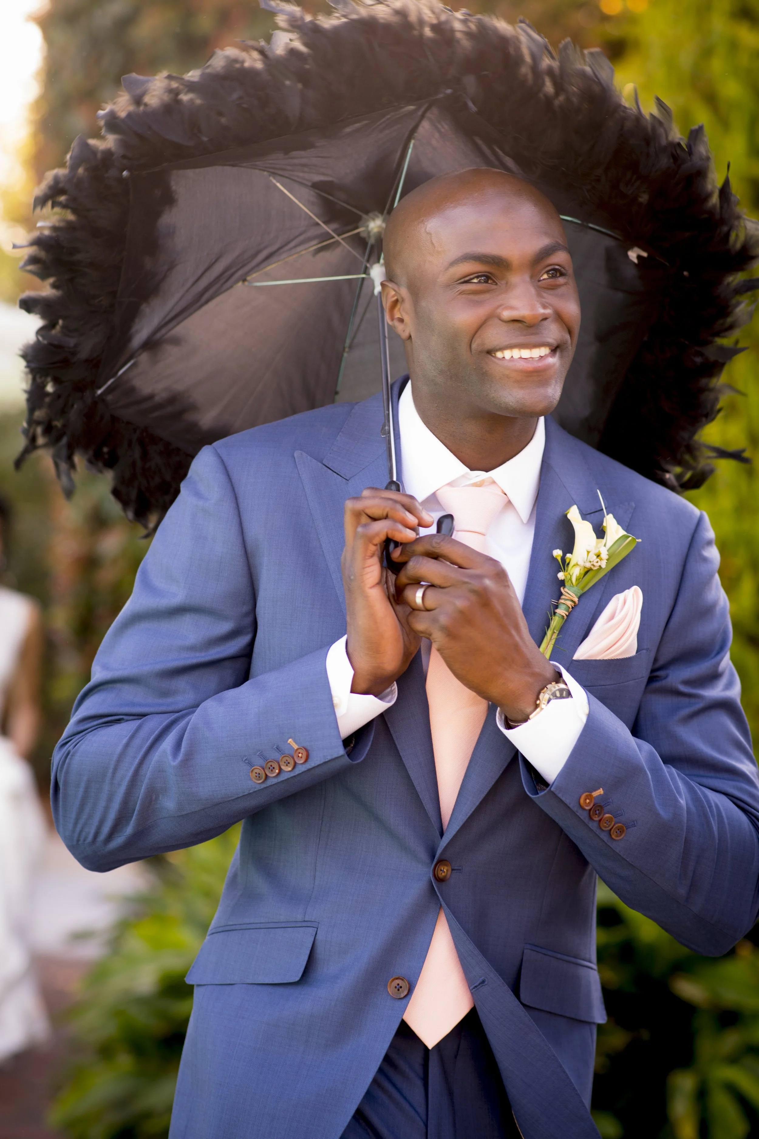 A smiling man with dark skin and a shaved head, dressed in a blue suit, holding a large black feathered umbrella, outdoors with blurred greenery in the background.