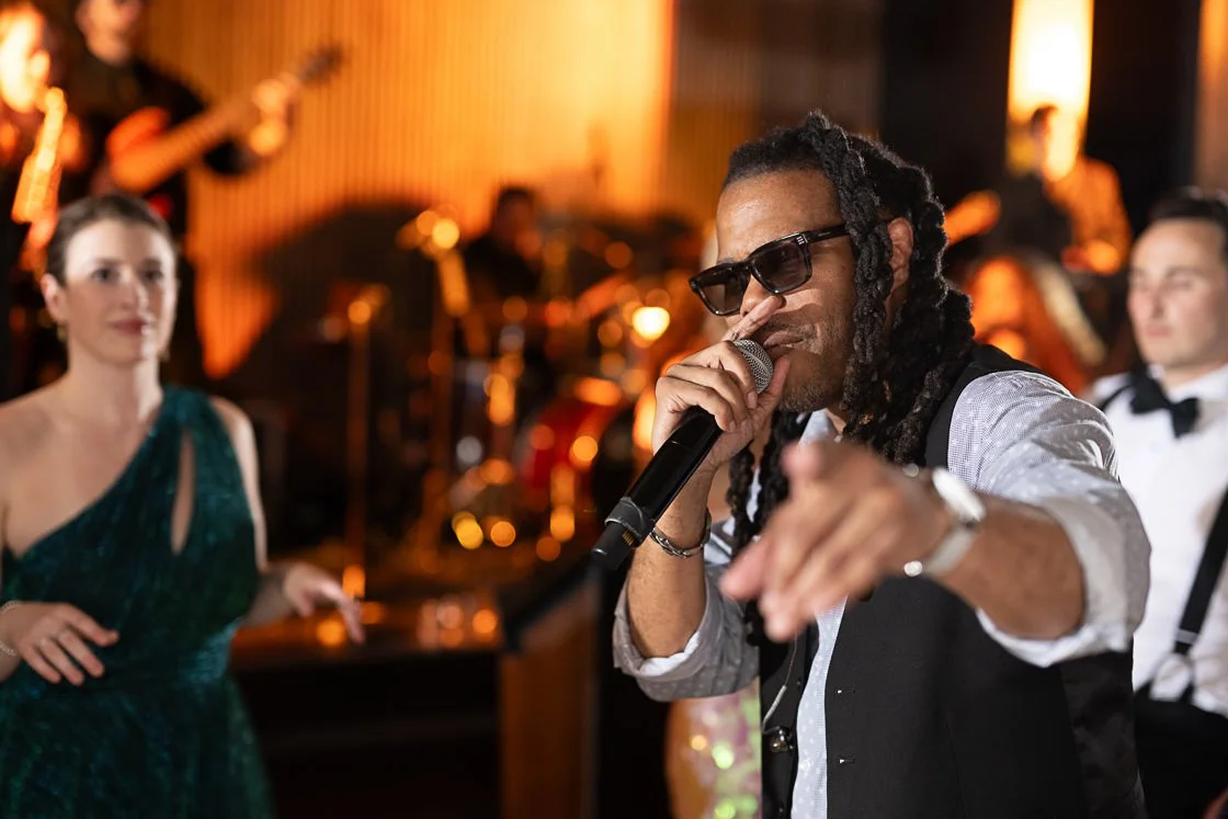 A singer with dreadlocks, sunglasses, and a vest performs with a microphone, pointing toward the camera at a party or concert. A woman in a green dress and other musicians are visible in the background.