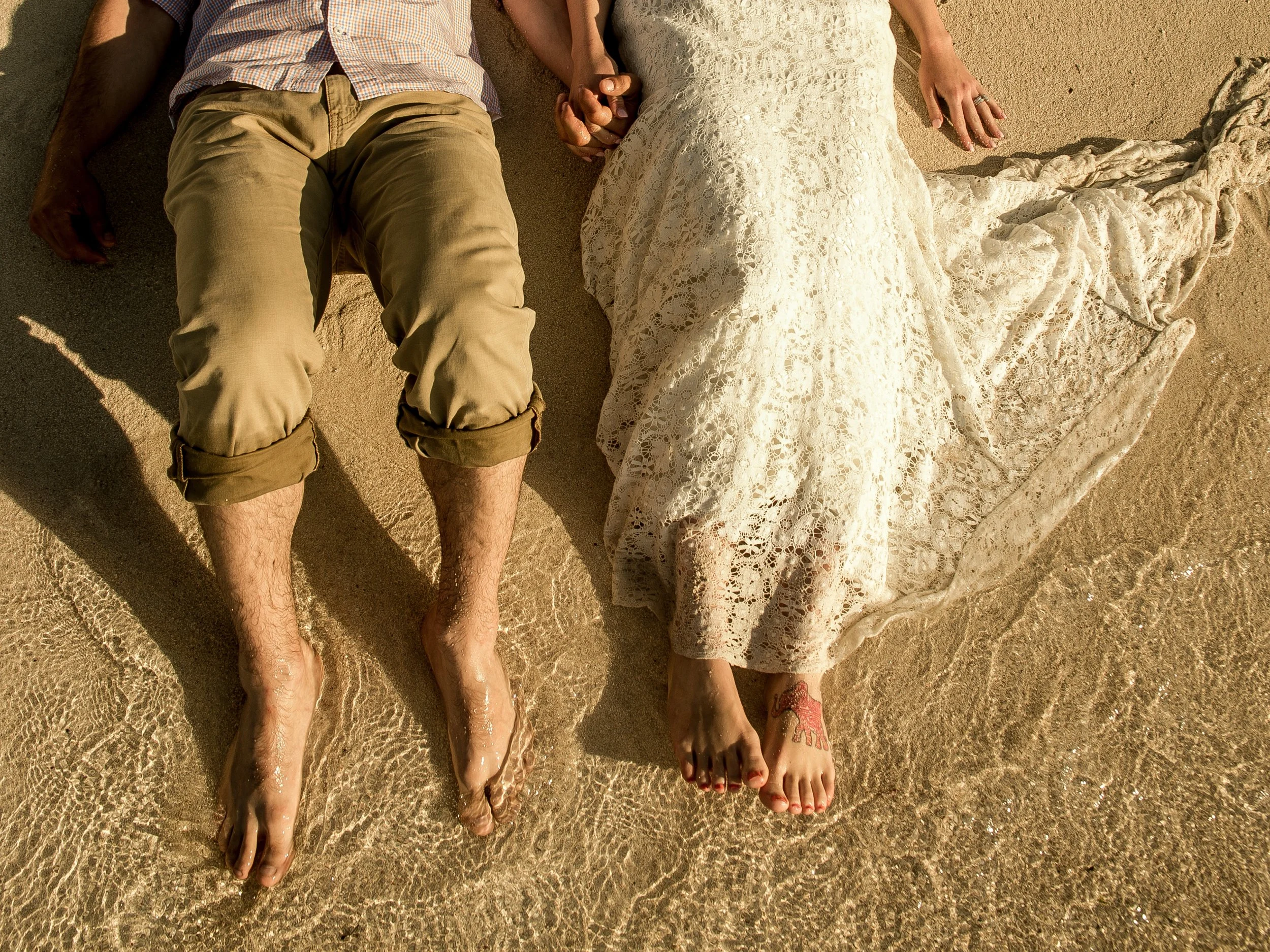 A couple lying on the sand at the beach, holding hands. Only their legs, partly their feet, and hands are visible. One is wearing light brown pants and a checkered shirt, the other is wearing a white lace dress. Their feet are in the shallow water wi