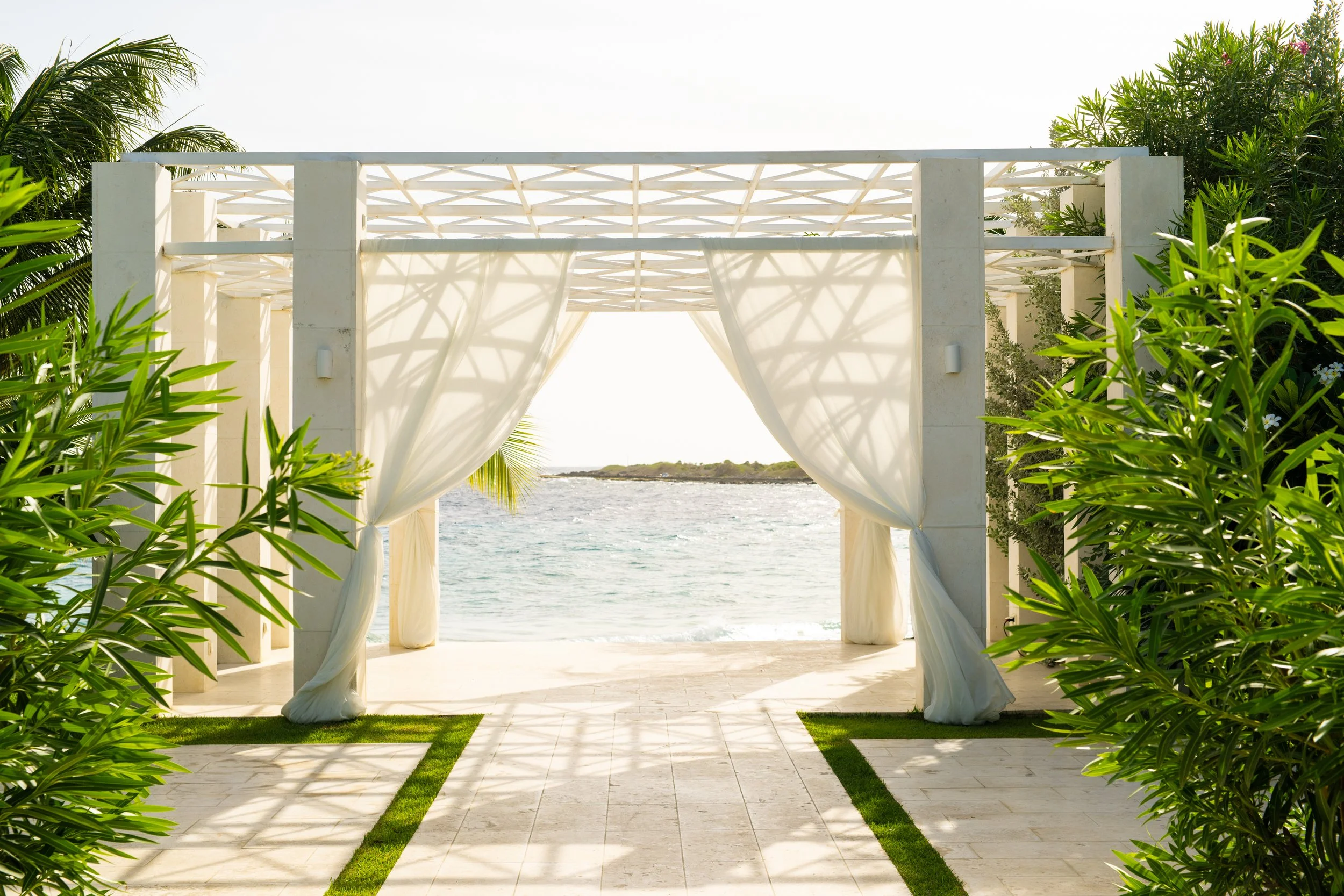 Beach wedding arch with white curtains, overlooking the ocean, framed by green plants.