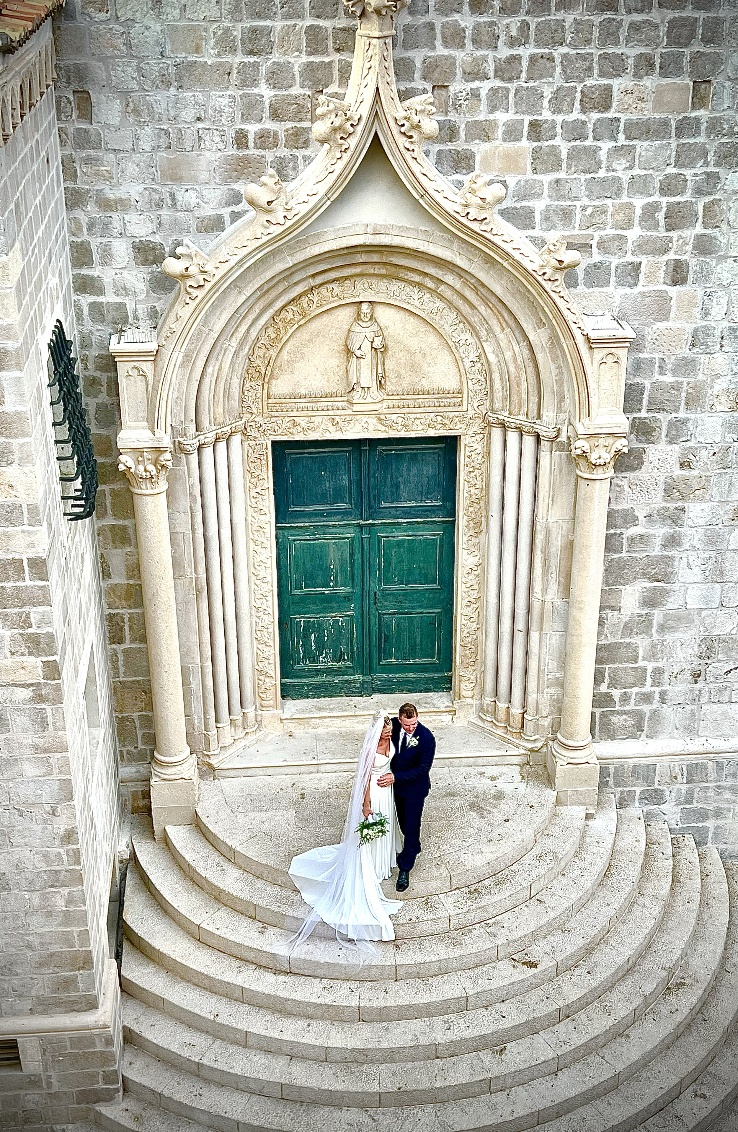 A bride and groom standing on the steps of a church in front of a large ornate doorway, holding each other and sharing a kiss. The bride is wearing a white gown and veil, holding a bouquet, while the groom is in a dark suit. The church has a stone fa