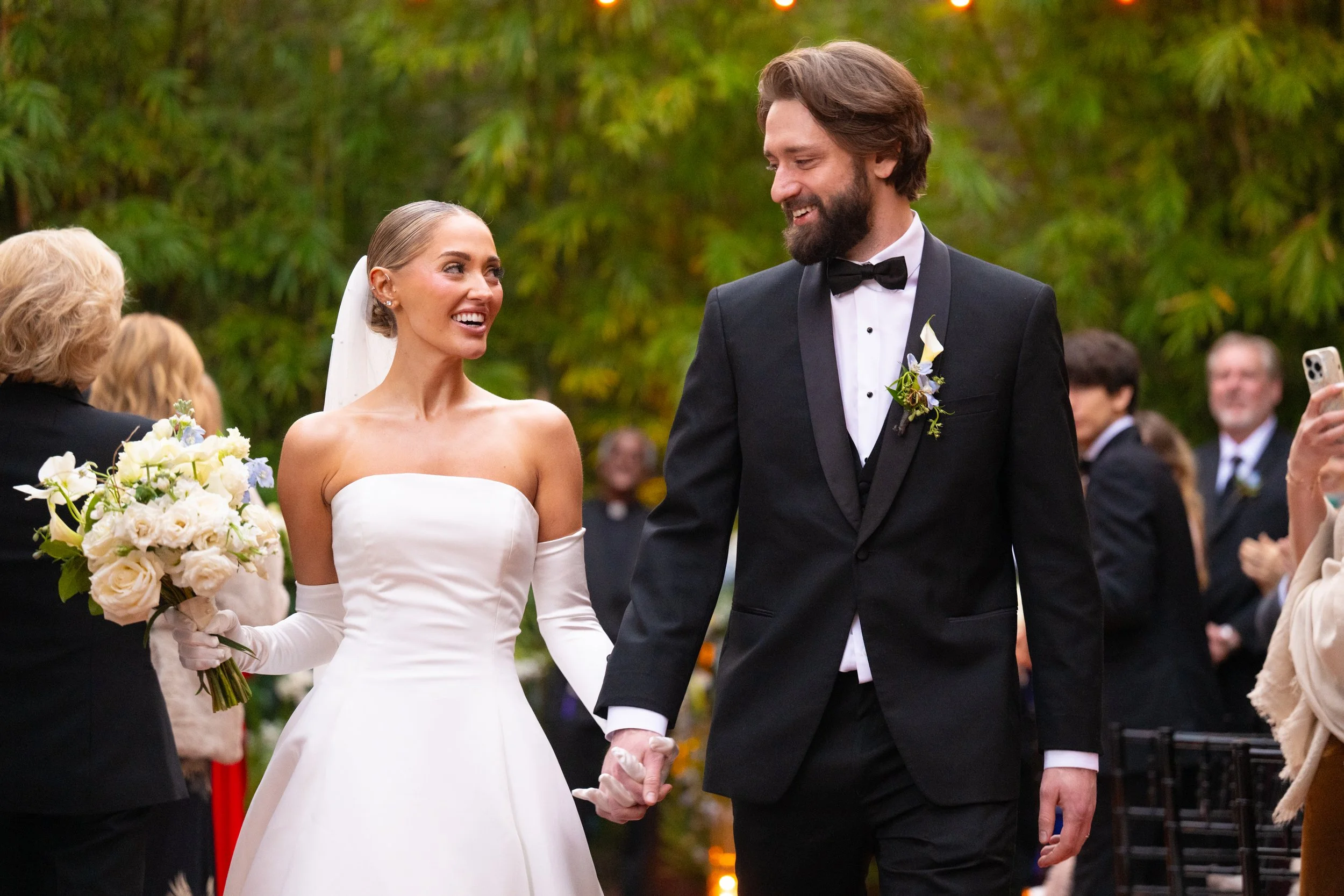 A bride and groom holding hands during their wedding ceremony outdoors, with guests in the background.