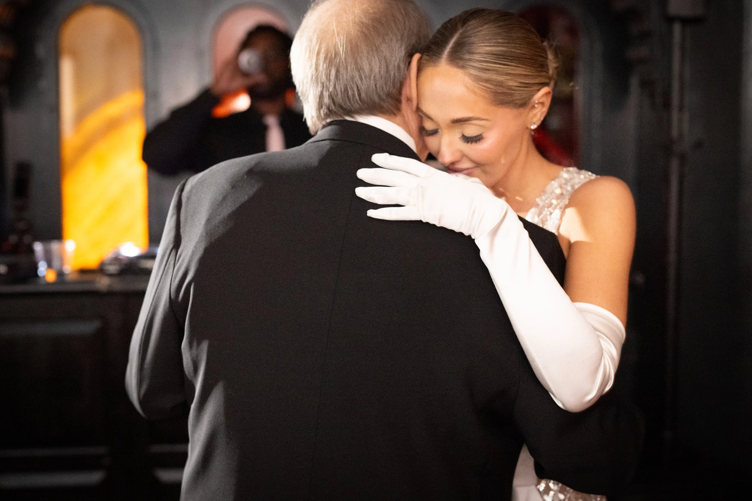 A man and woman dancing closely at a wedding reception, with the man wearing a black suit and the woman wearing an elegant dress with white gloves.