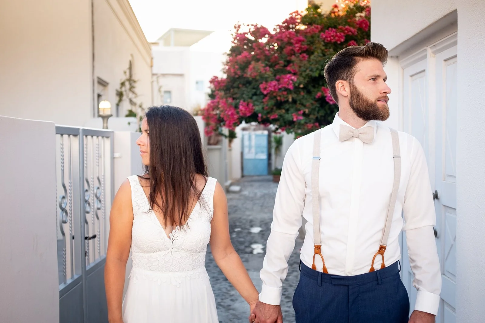 A couple dressed in wedding attire holding hands outdoors near white walls and a flowering pink tree.