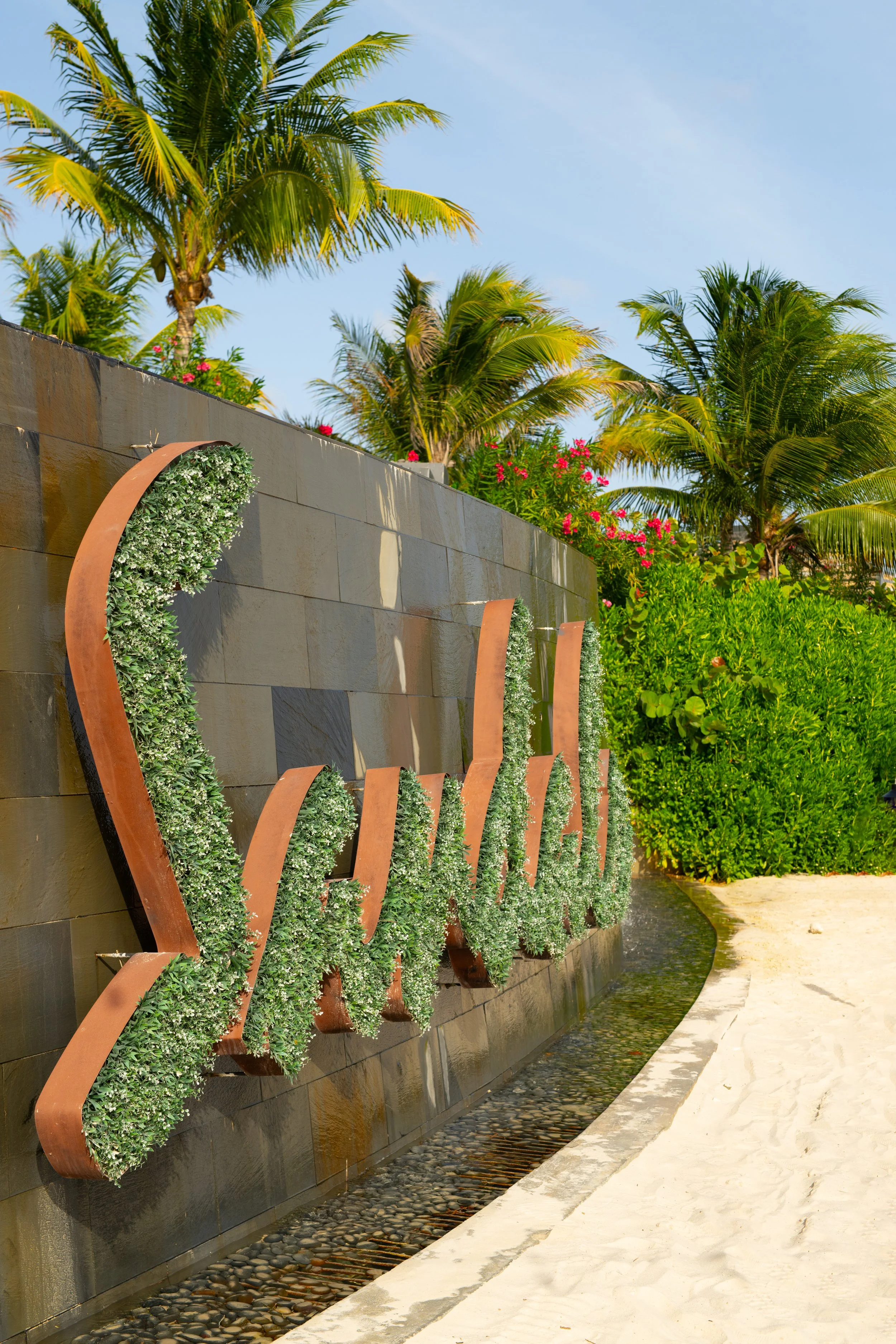 A curved stone wall with metal and plant decorations, sunlit palm trees, and bright pink flowers in a tropical garden setting.