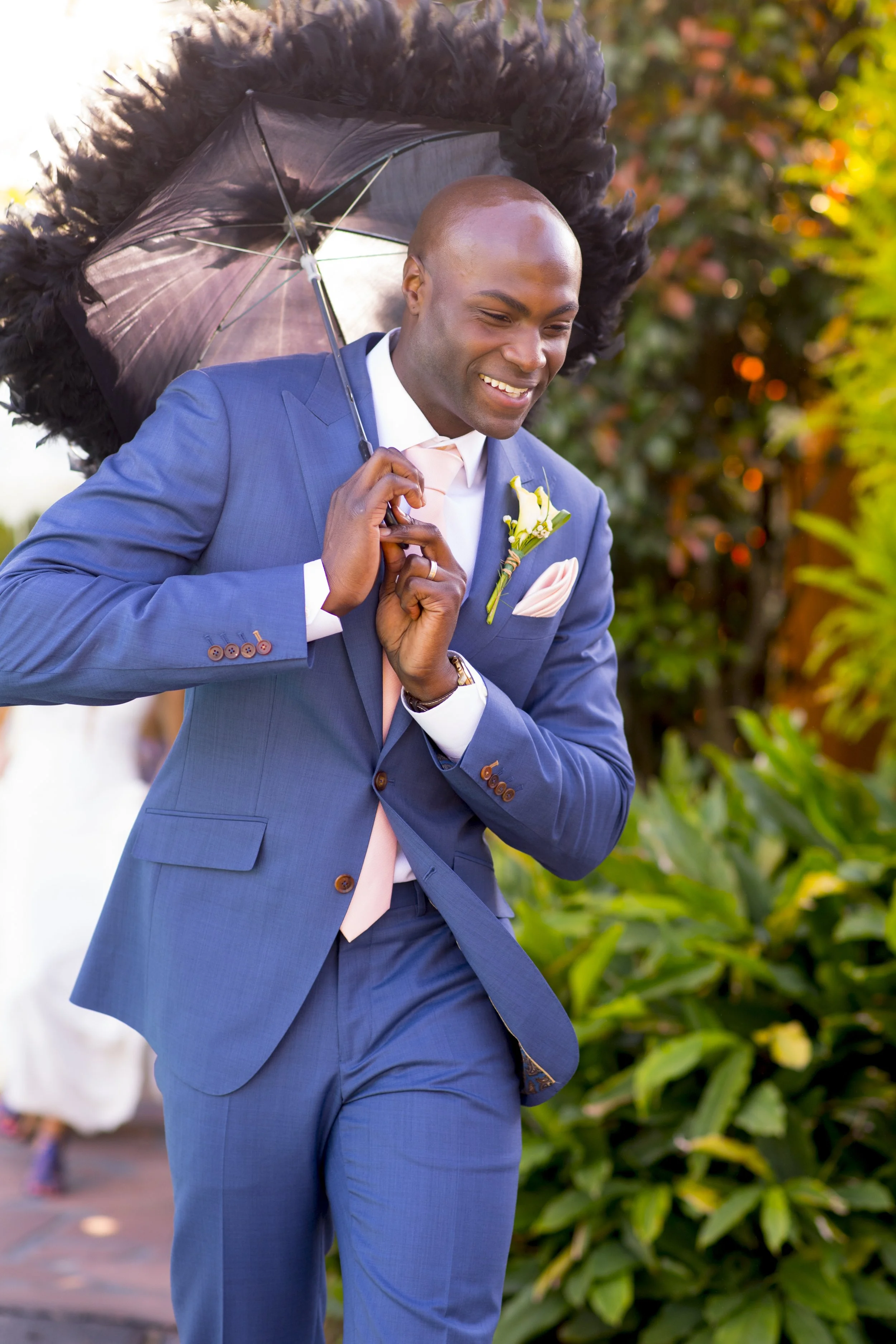 A man wearing a blue suit with a white shirt and light pink tie, holding a black umbrella with black feathers around the edge, smiling as he walks outdoors on a sunny day with green and orange foliage in the background.