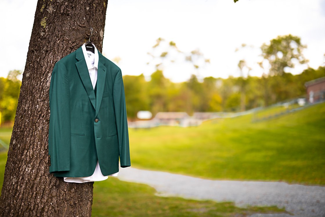 A green suit jacket hanging on a tree trunk with a white shirt underneath, in an outdoor park setting.