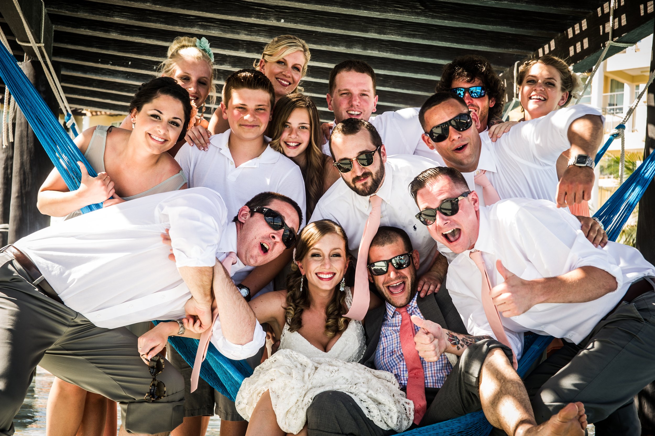 Group of people celebrating and taking a group photo under a wooden structure, some wearing sunglasses, smiling, and dressed in summer clothing.