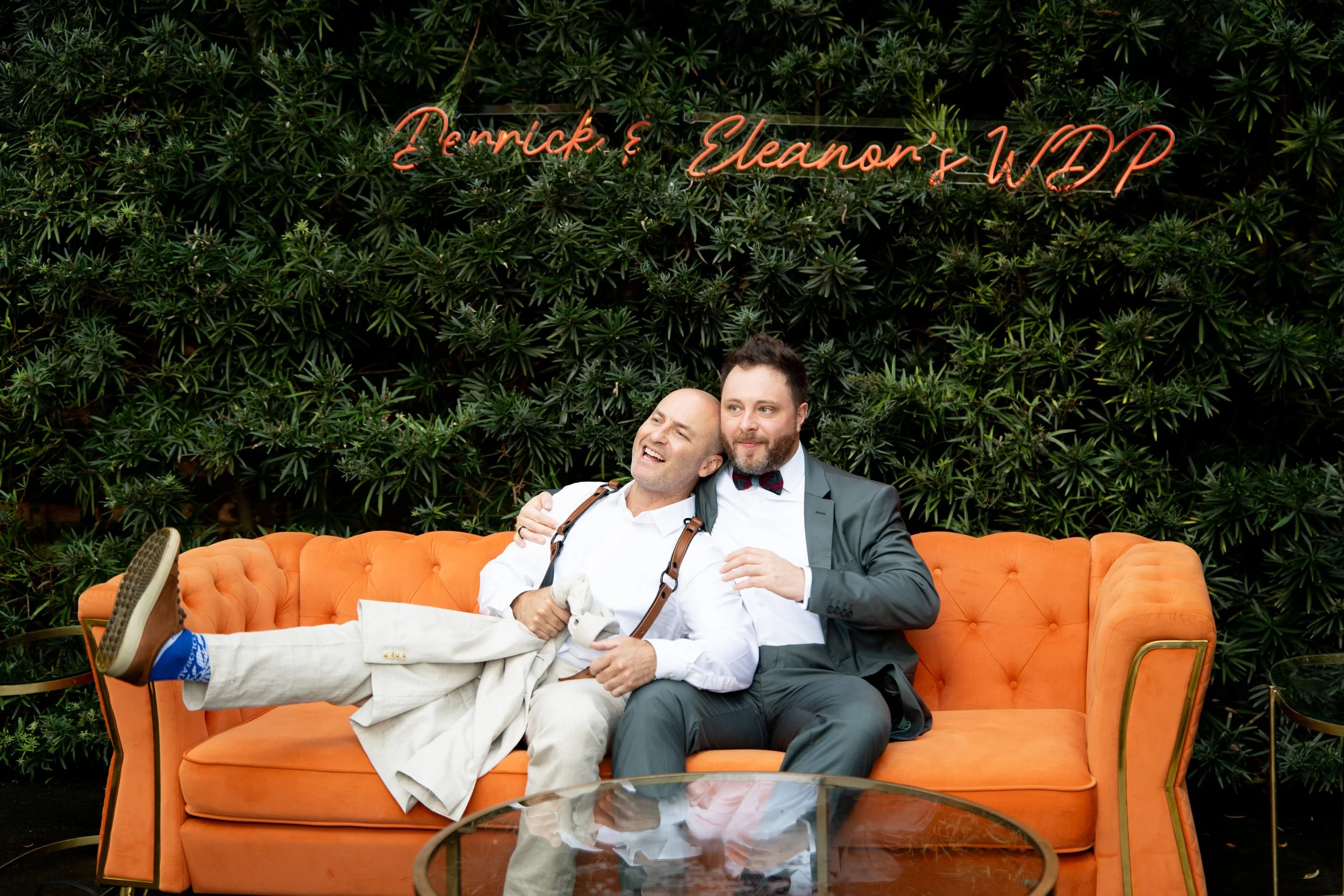 Two men sitting on an orange couch in front of a green foliage wall with a neon sign that reads 'Terrick & Eleanor's WED' and a glass coffee table in the foreground.