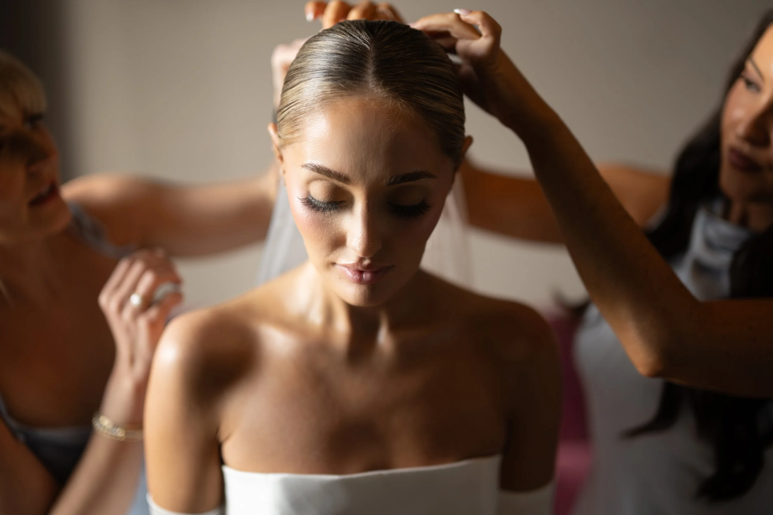 A bride with her eyes closed, having her hair styled by two women on either side, in a room with soft lighting.