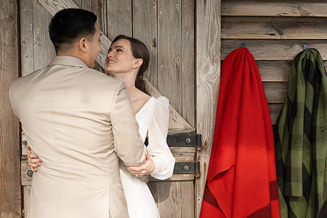 A couple dressed in wedding attire, smiling and gazing at each other, standing close to a rustic wooden wall with red and green blankets hanging on hooks.