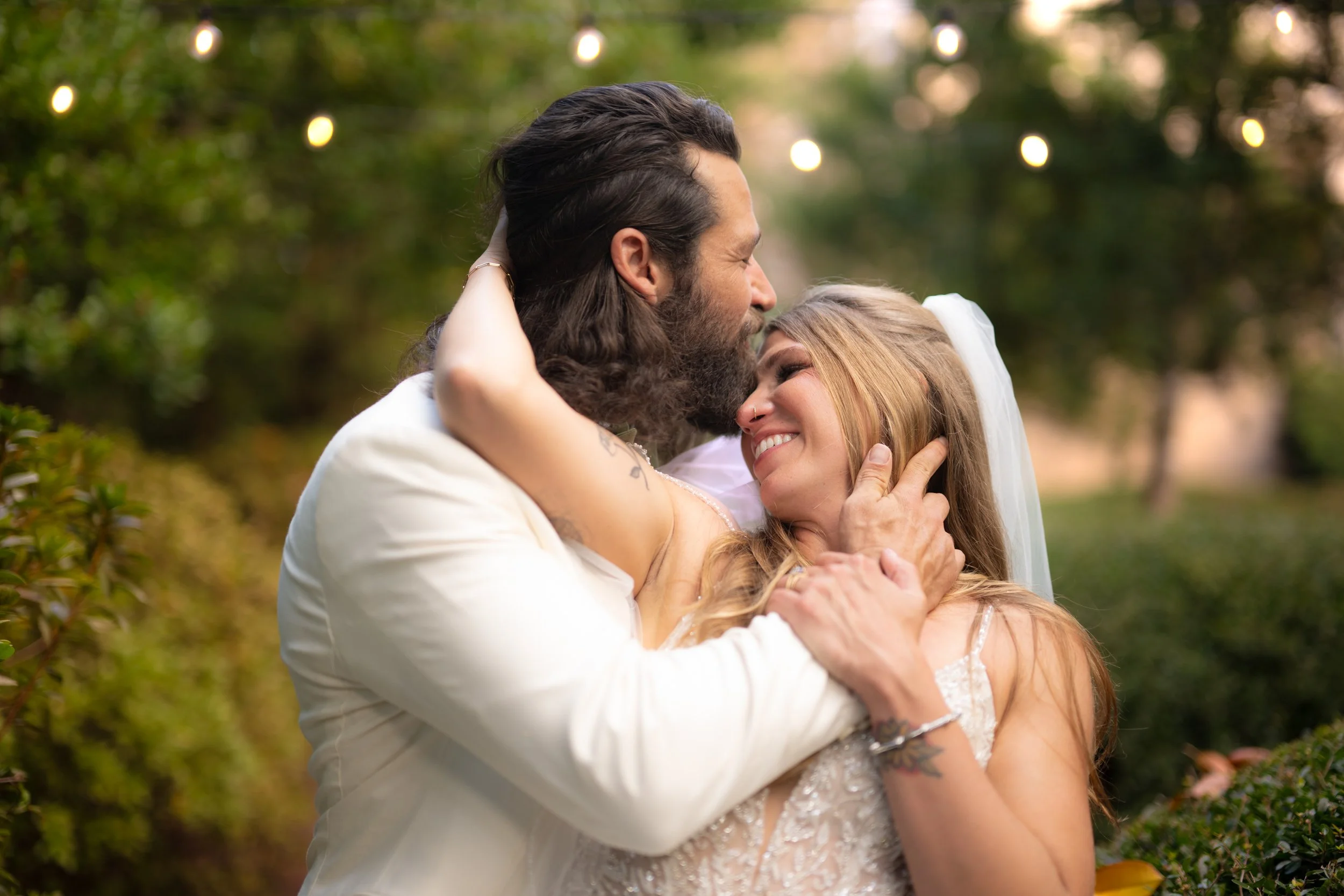 A couple embraces during their wedding ceremony outdoors, surrounded by greenery and warm string lights.