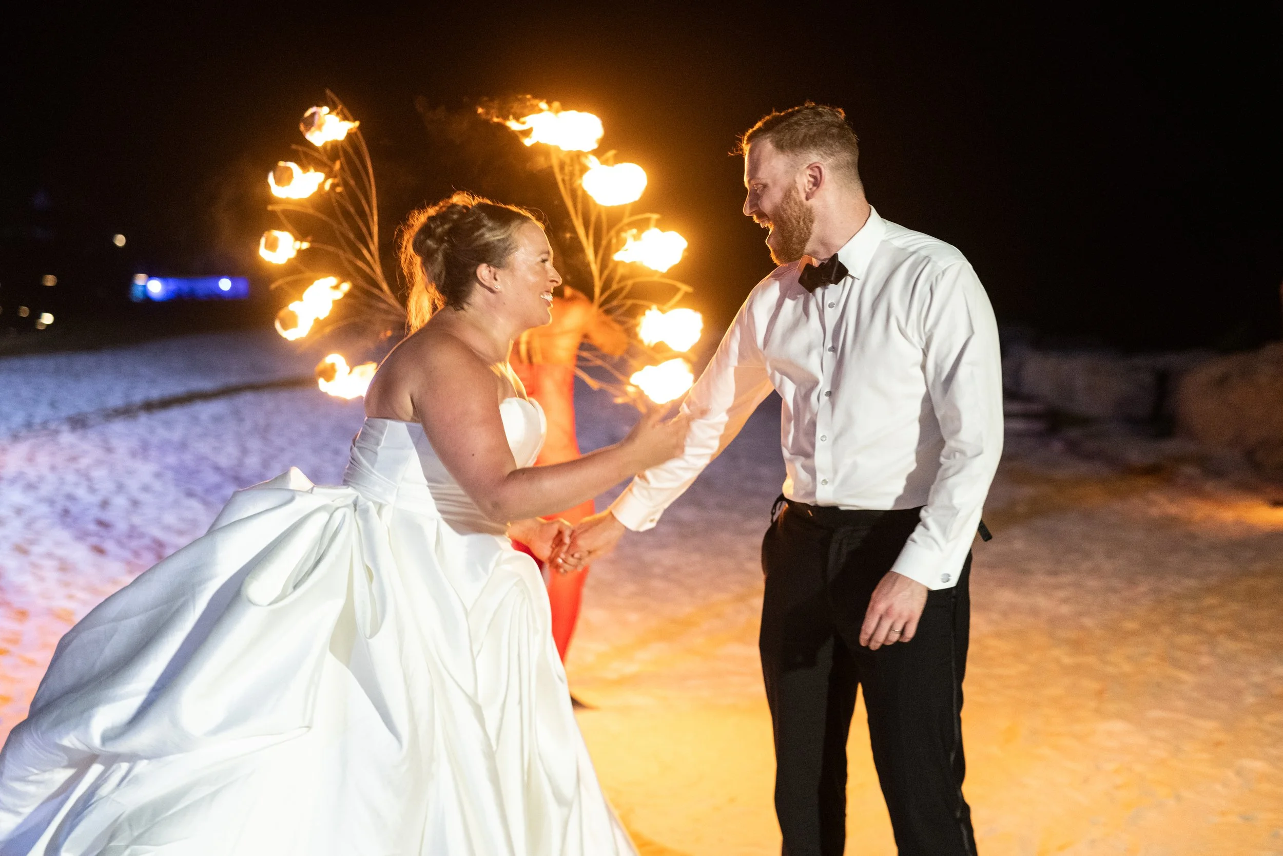 A bride and groom dancing at their wedding reception on a beach at night, with fire torches in the background.