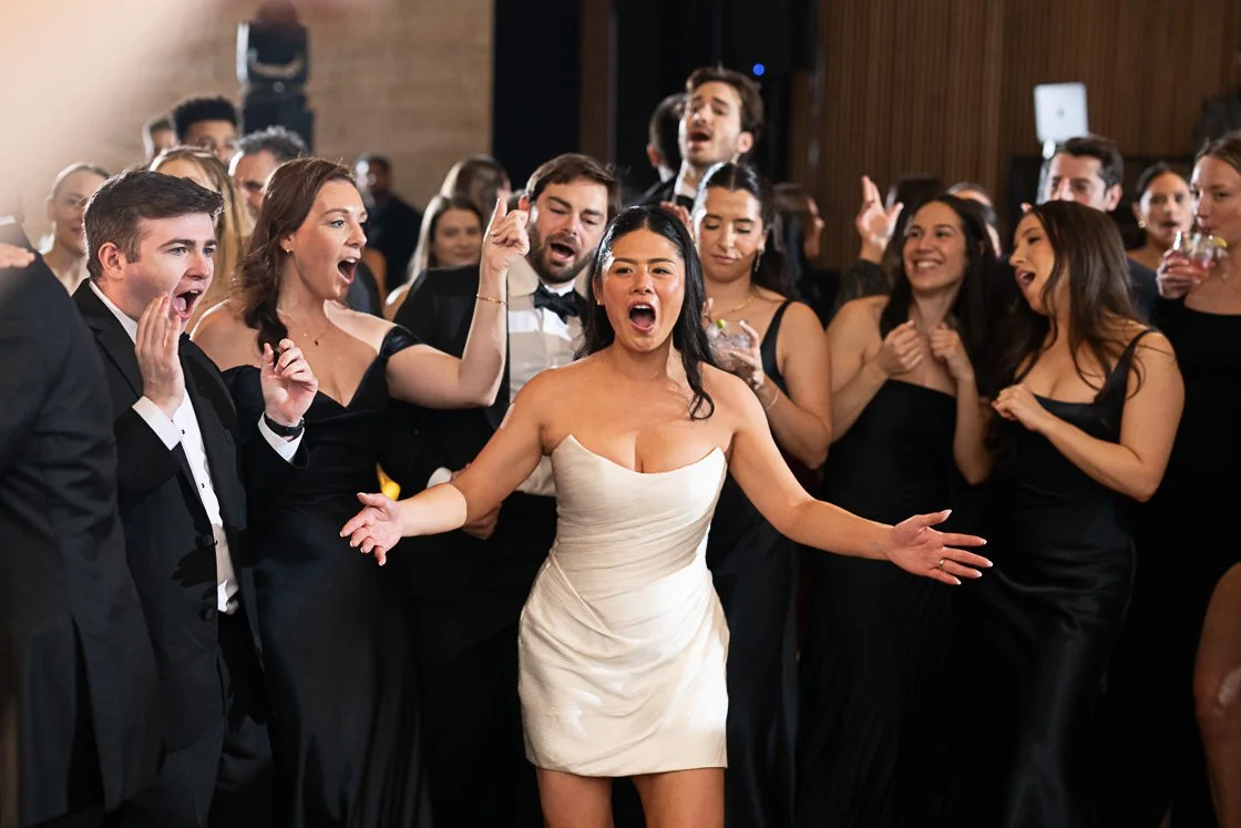 A woman in a white dress dancing and singing with a group of people at a celebration or wedding reception.