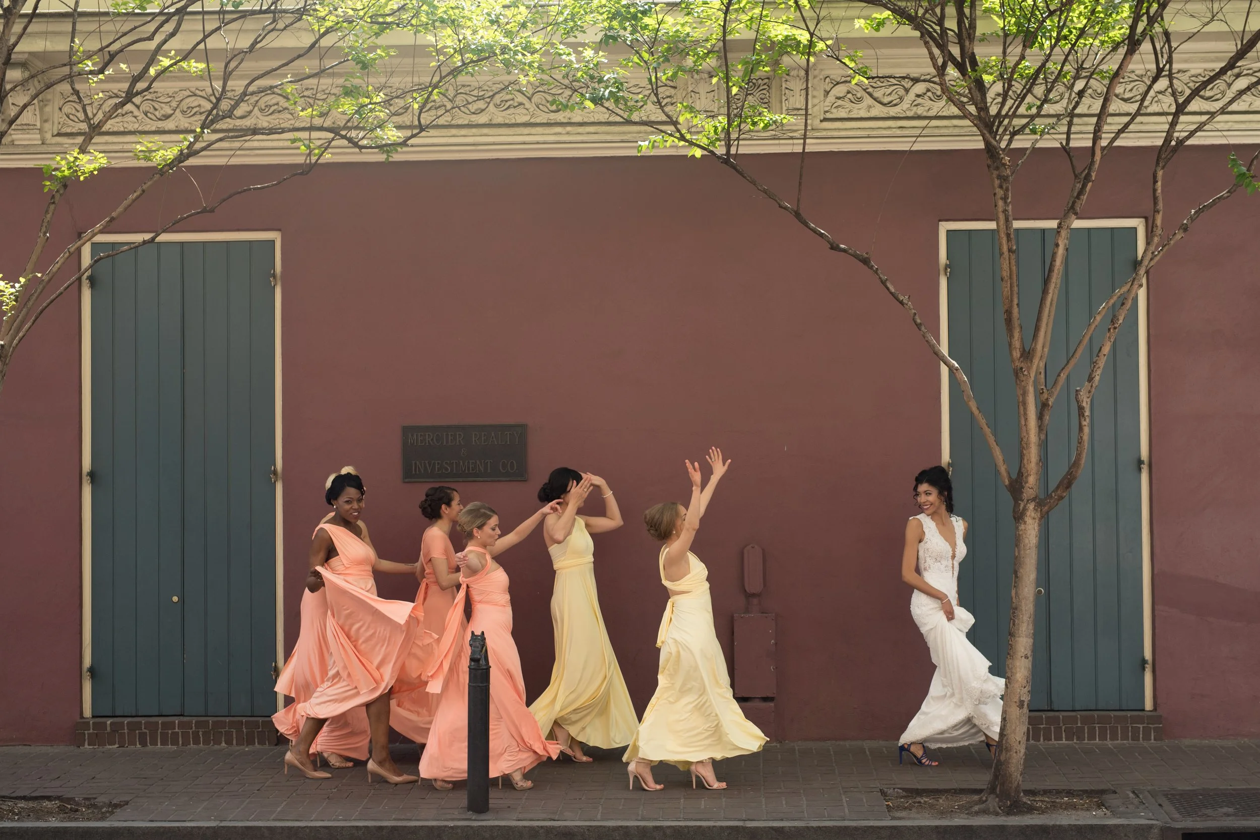 A group of women in colorful dresses are walking along a sidewalk in front of a building with green shutters and a maroon wall. There is a tree in the foreground, and a sign that reads 'Mercier Realty & Investment Co.'