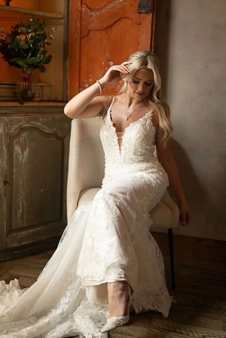 A bride in a white lace wedding dress sitting on a chair, adjusting her hair in a rustic room with wooden furniture and a vase of flowers in the background.