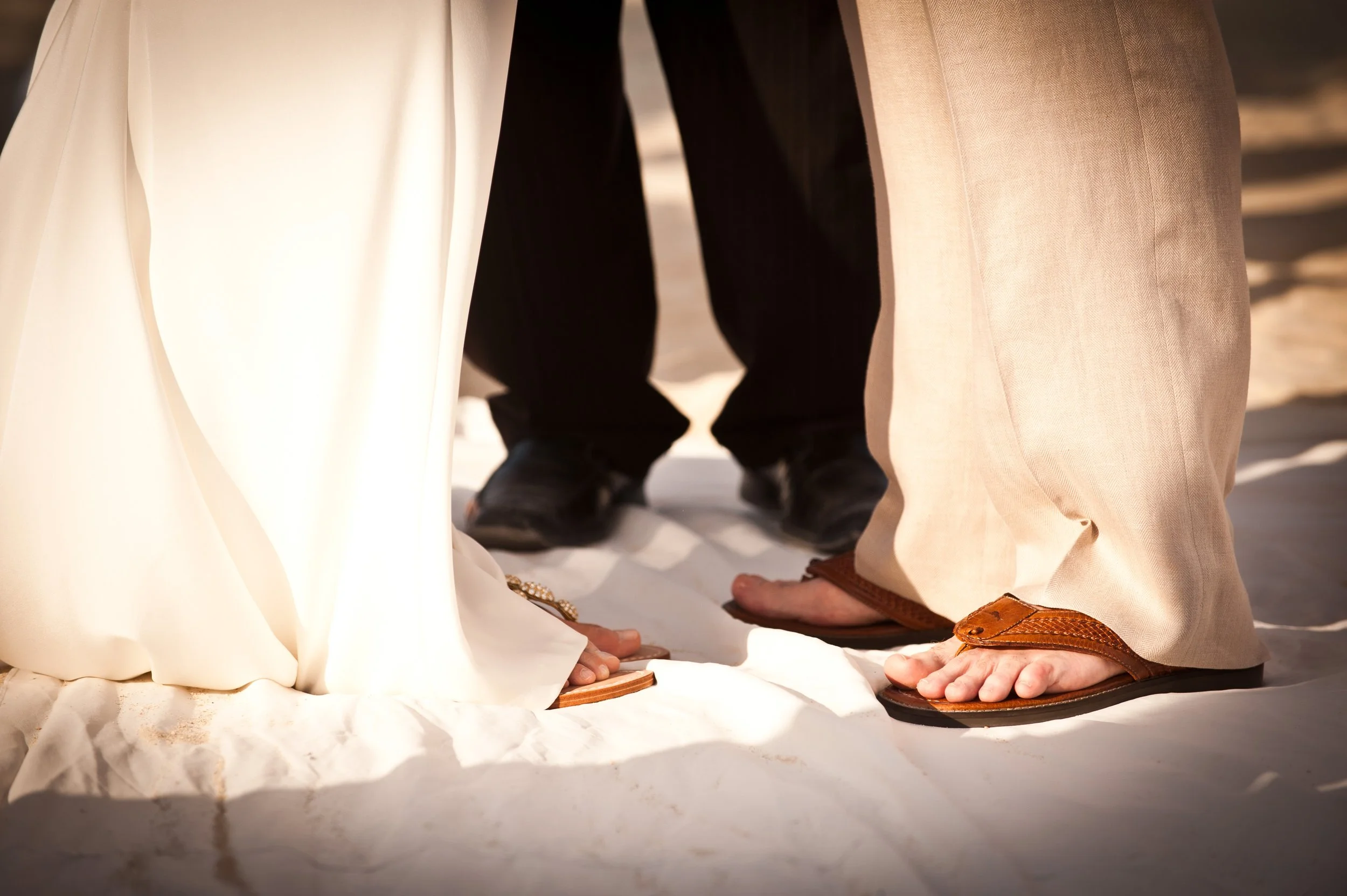 Close-up of feet and lower legs of people dressed in traditional, formal or ceremonial attire, standing on a white surface with some white fabric and shadows visible.