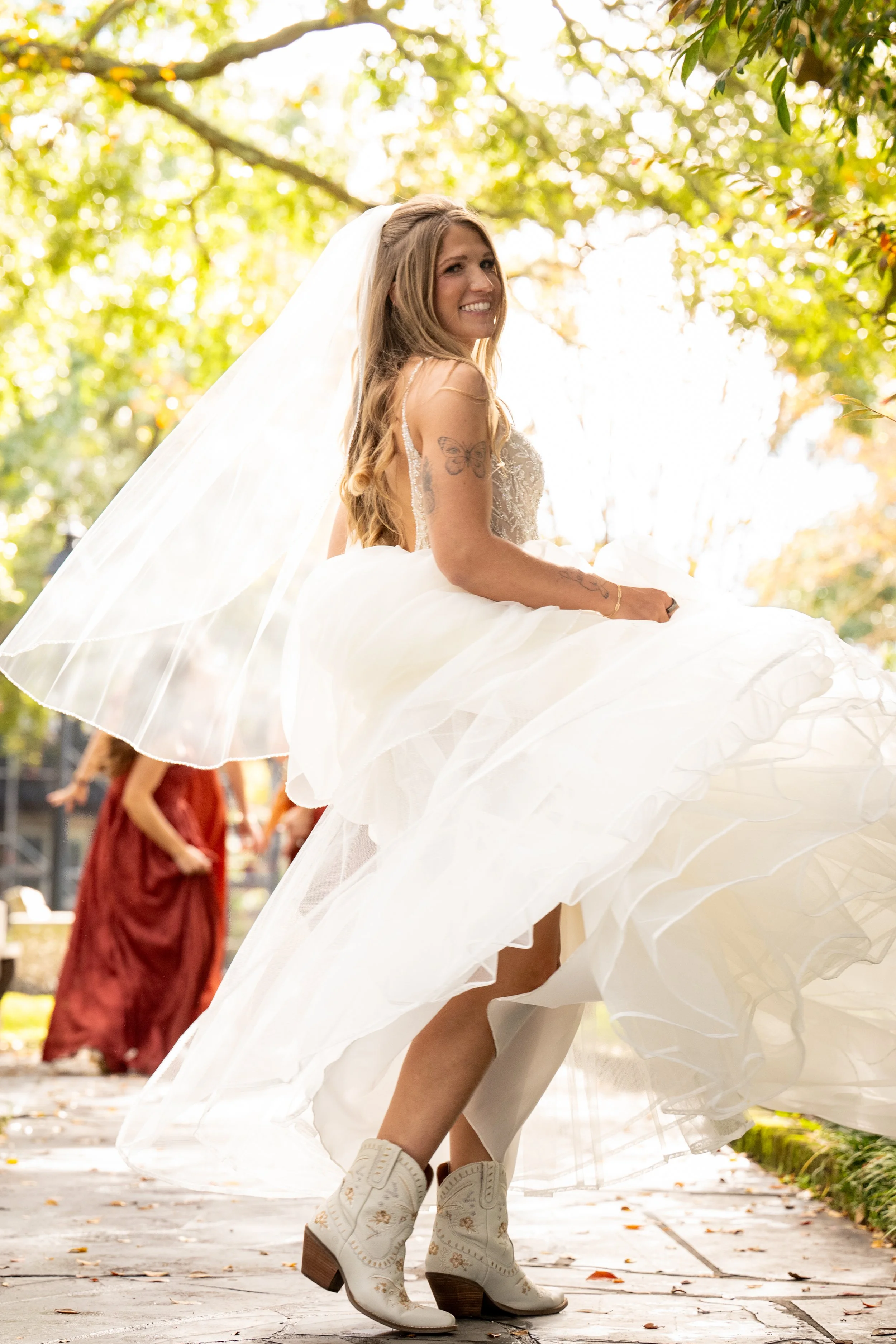 A bride in a white dress and cowboy boots twirls outdoors, with sunlight filtering through the trees and a woman in a maroon dress in the background.