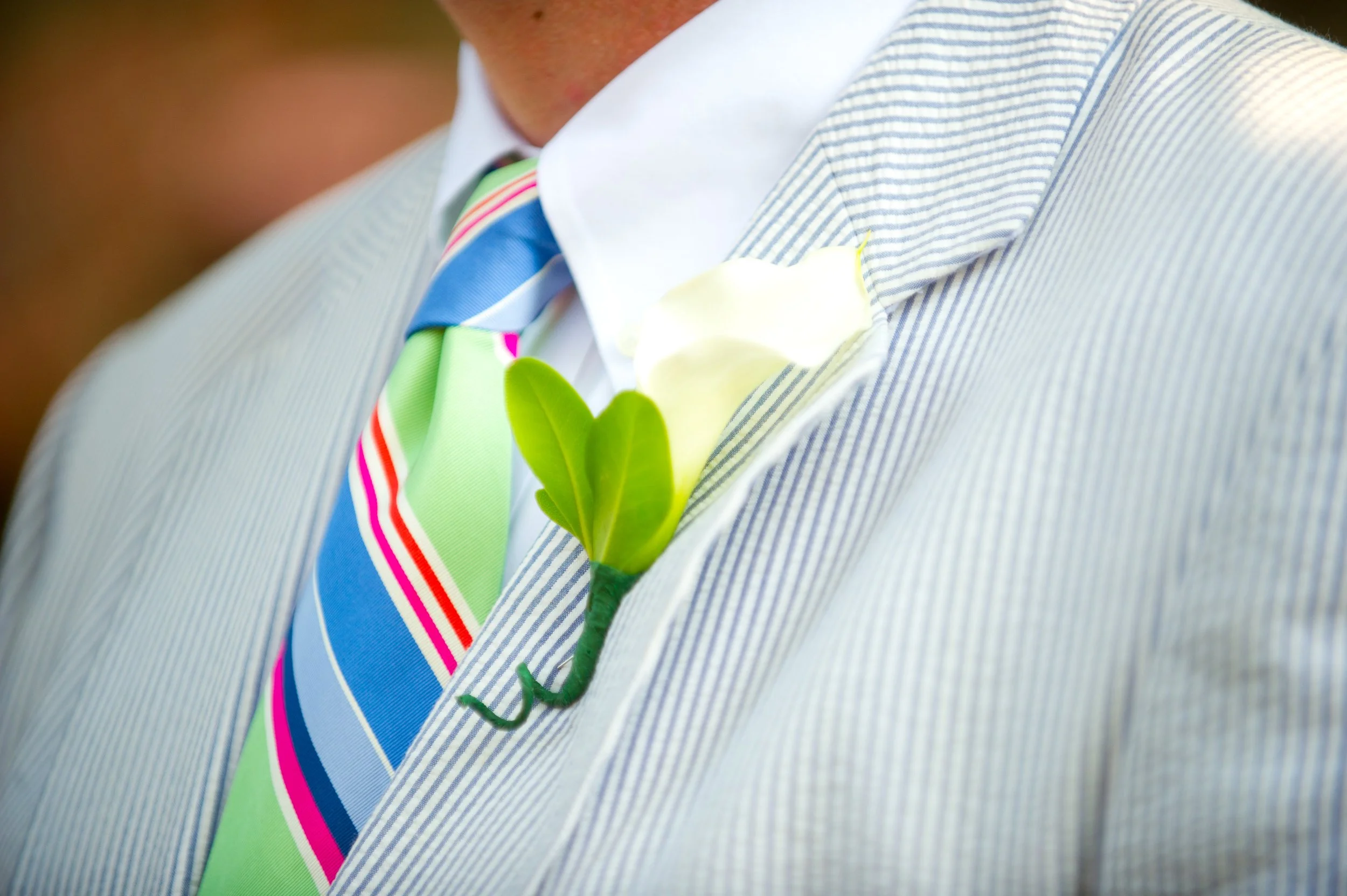 Close-up of a man wearing a striped suit, white shirt, colorful striped tie, and a boutonniere with green leaves.