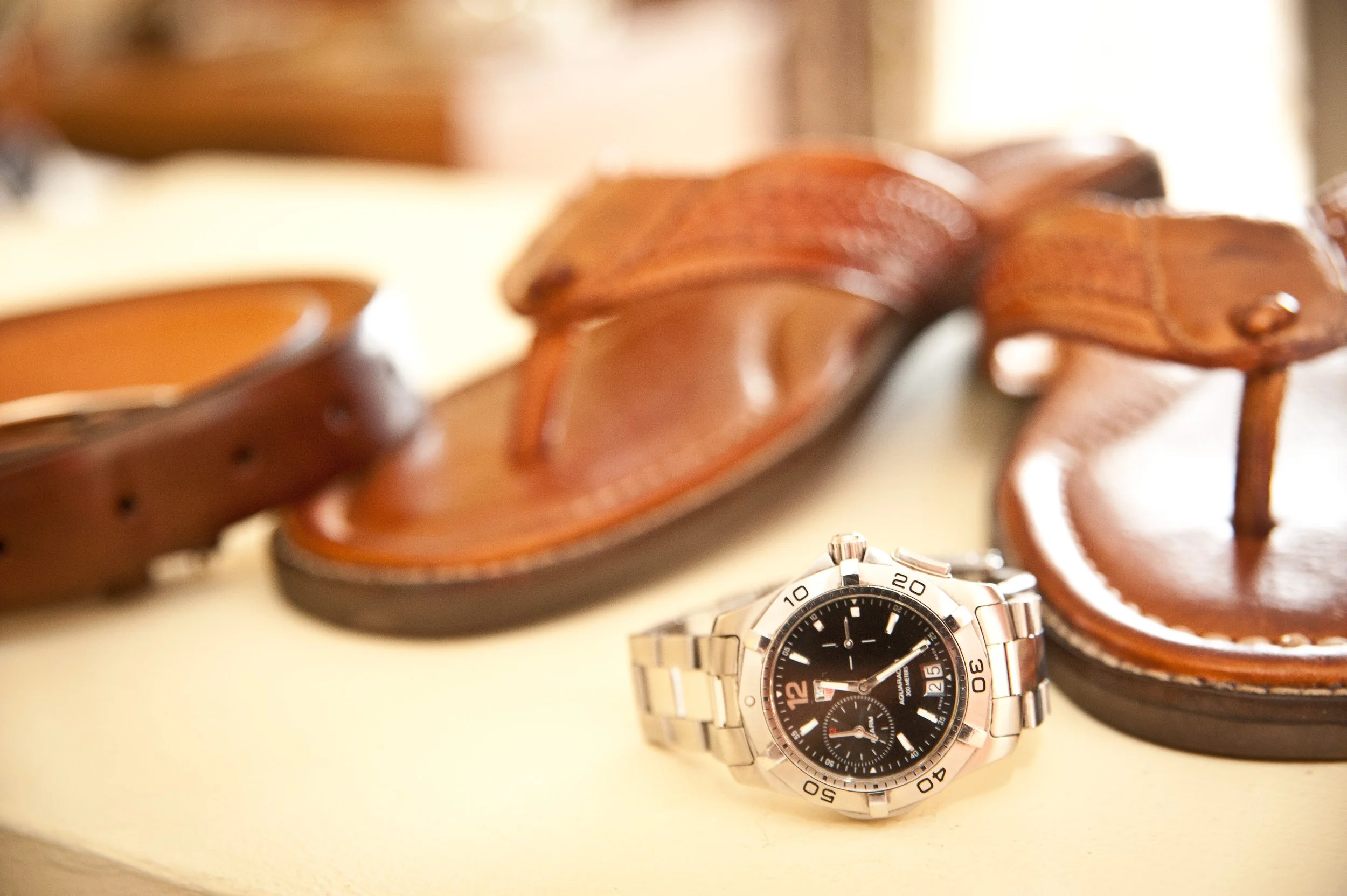 Close-up of a silver wristwatch with a black dial placed in front of a pair of brown leather shoes on a white surface.