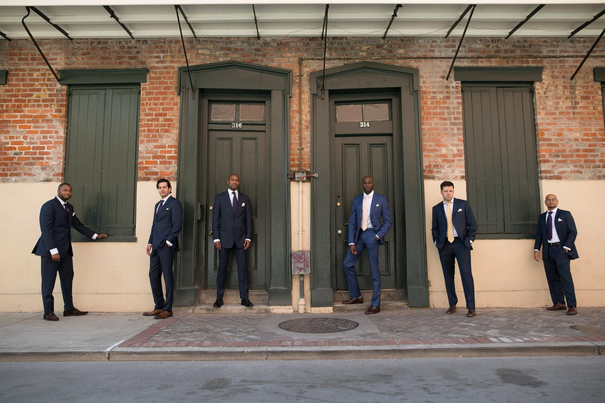 Six men dressed in suits are standing outside against a brick wall with three doors, two of which are closed and one partly open.