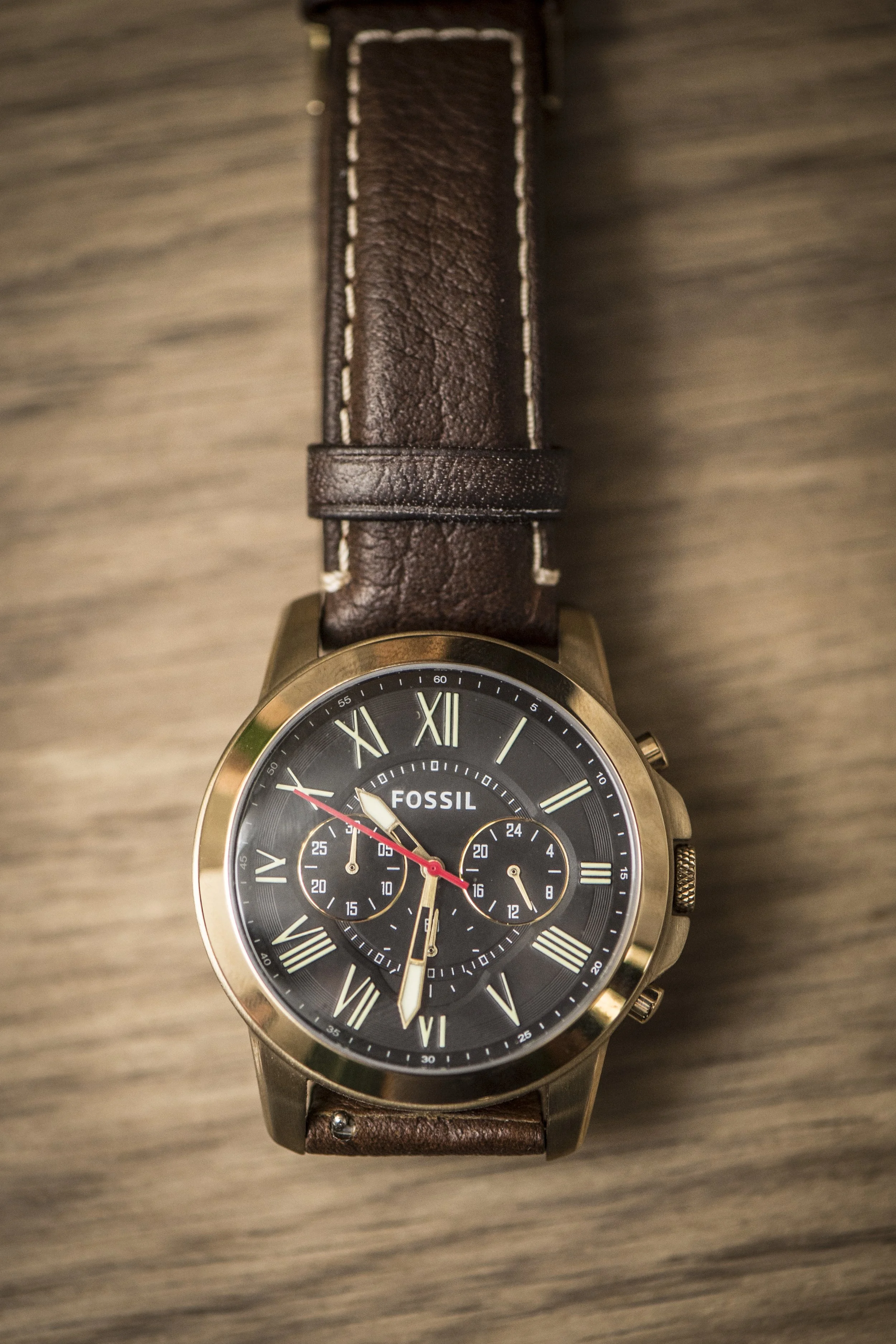 A Fossil wristwatch with a black dial, Roman numerals, three subdials, and a brown leather strap, placed on a wooden surface.