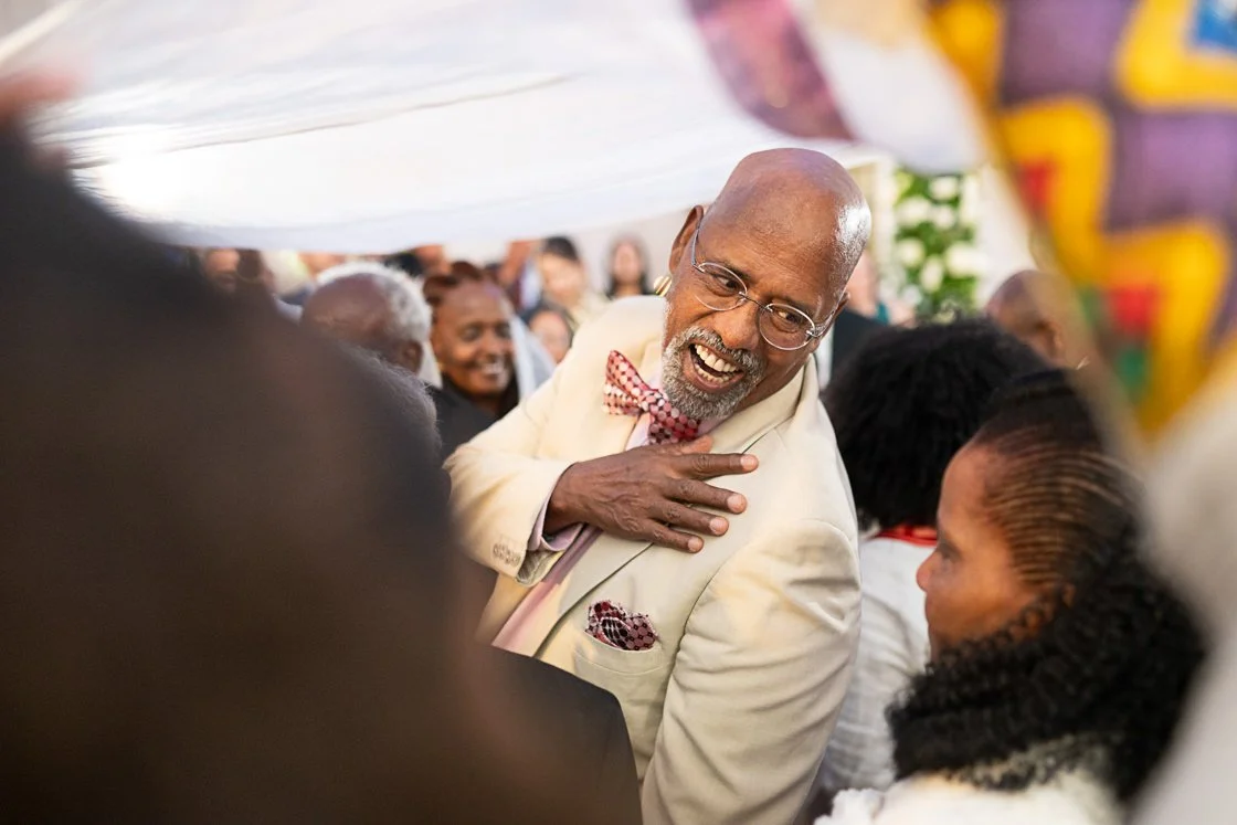 An older man with glasses, a gray beard, and a bald head, wearing a light-colored suit with a red bow tie, smiling and touching his chest while talking to a woman in a patterned dress during a social gathering.