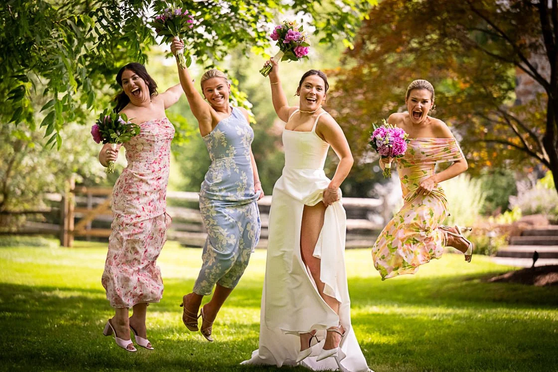 Four women in spring dresses celebrating outdoors, jumping with bouquets of flowers in their hands, with lush green trees and a fence in the background.