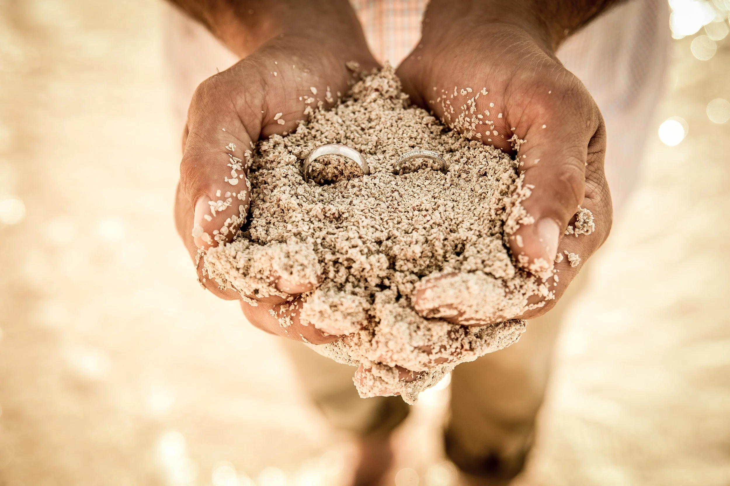 Close-up of a hand holding a handful of coarse, grainy sand, with two wedding rings partially embedded in the sand.
